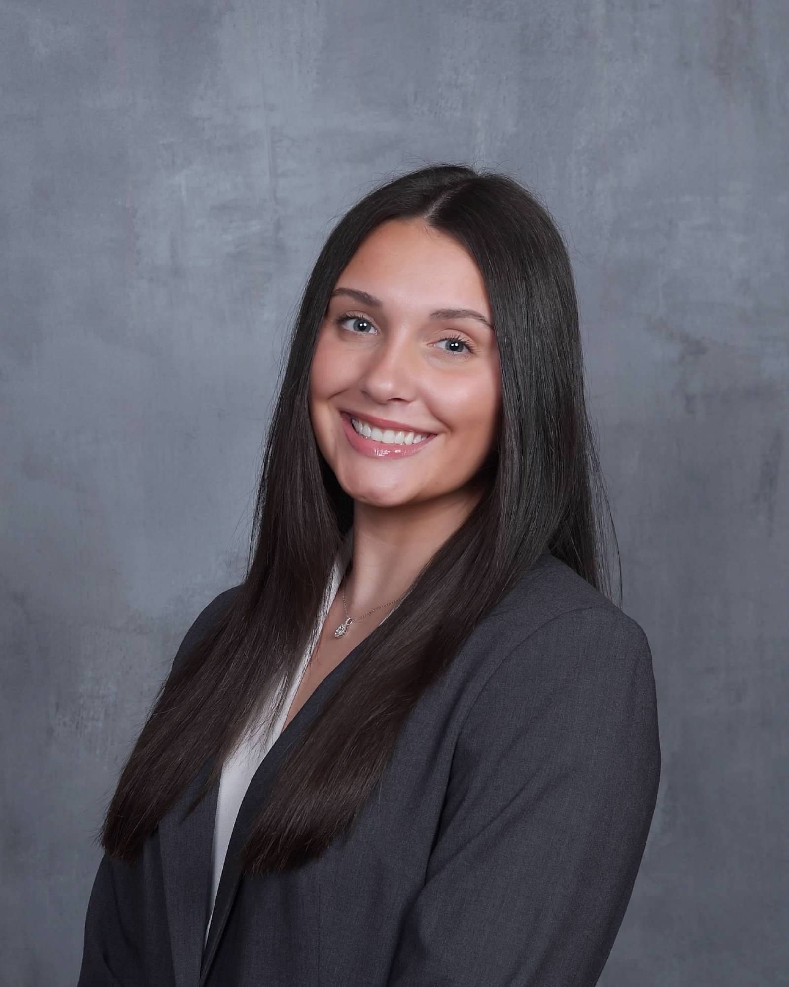 Smiling professional woman with long dark hair wearing a gray blazer and white blouse against a gray textured background.