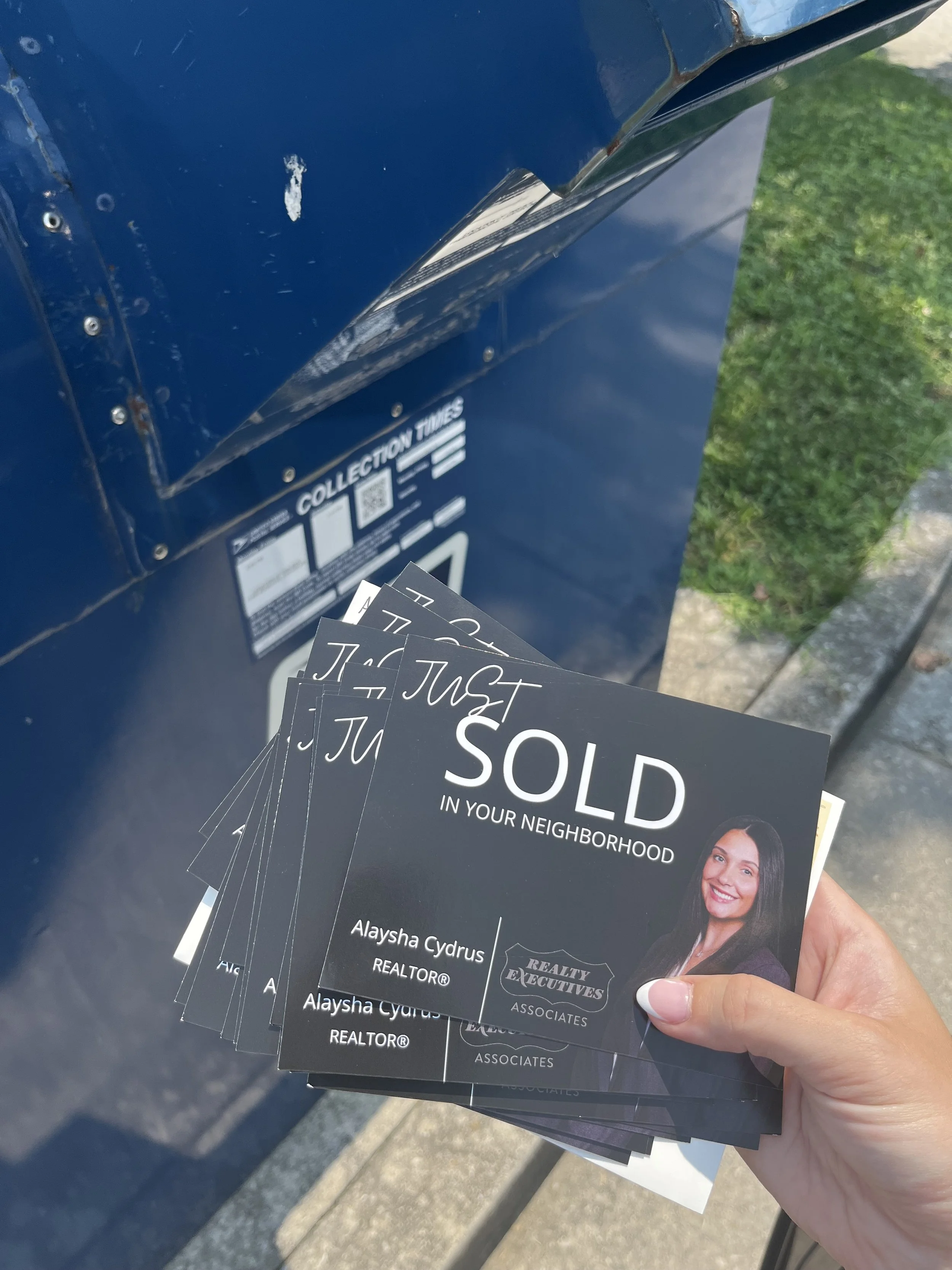 Hand holding black real estate postcards reading “Just Sold in Your Neighborhood” beside a blue mailbox.