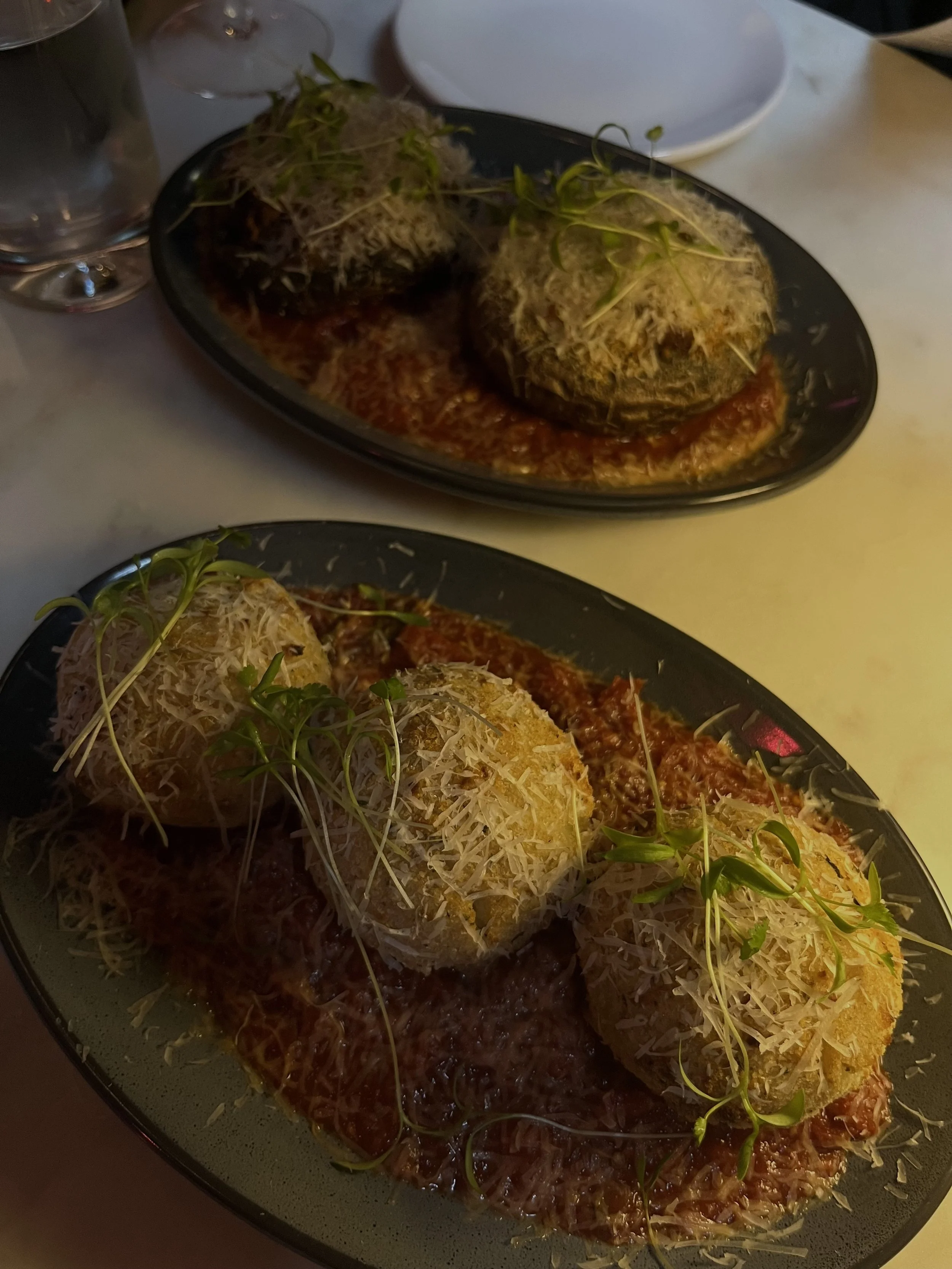 Two plates of saucy fried chicken cutlets topped with shredded cheese and microgreens.