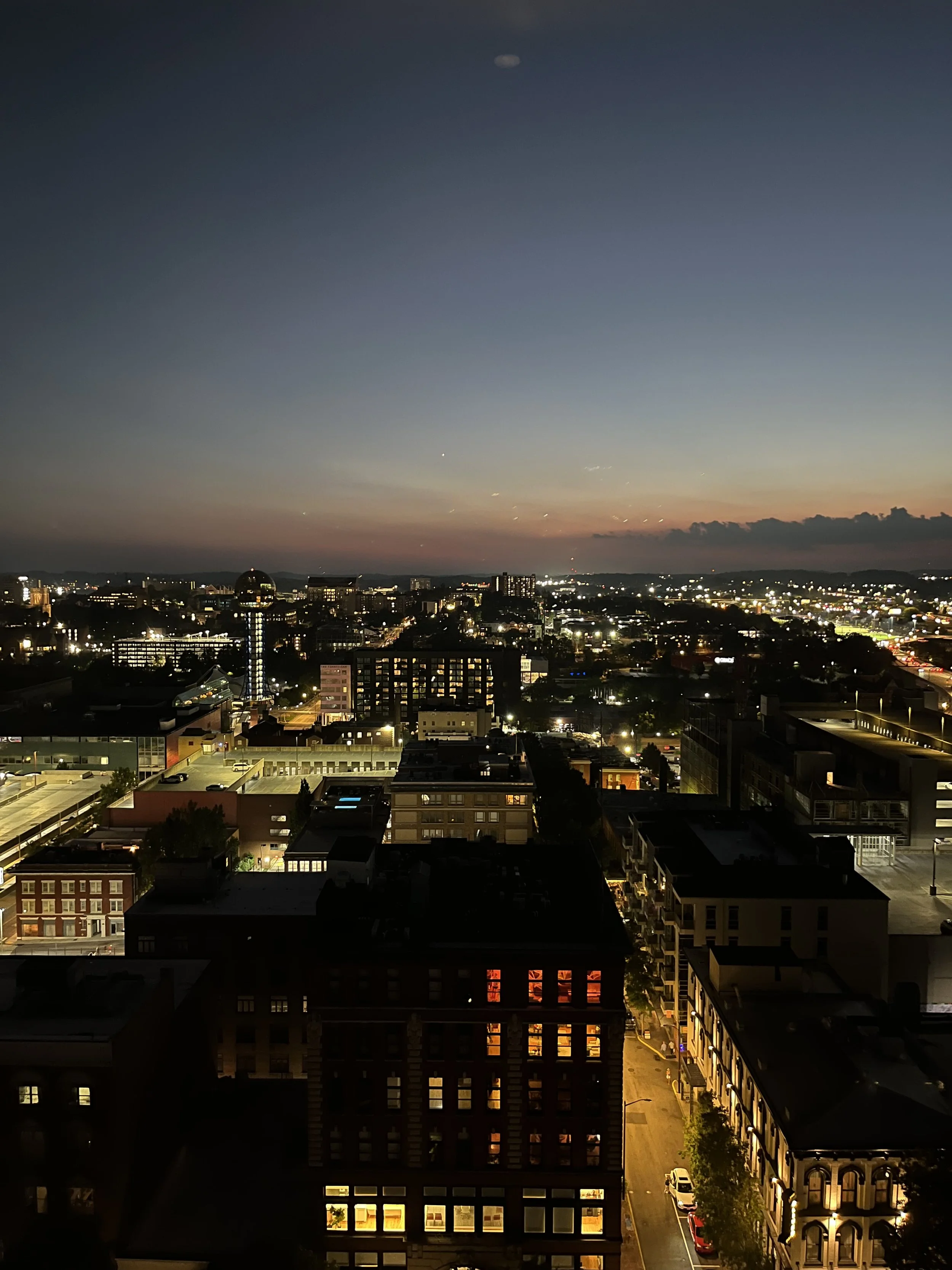 Twilight panoramic view of a brightly lit city skyline, downtown buildings, streets, distant hills, and clouds.