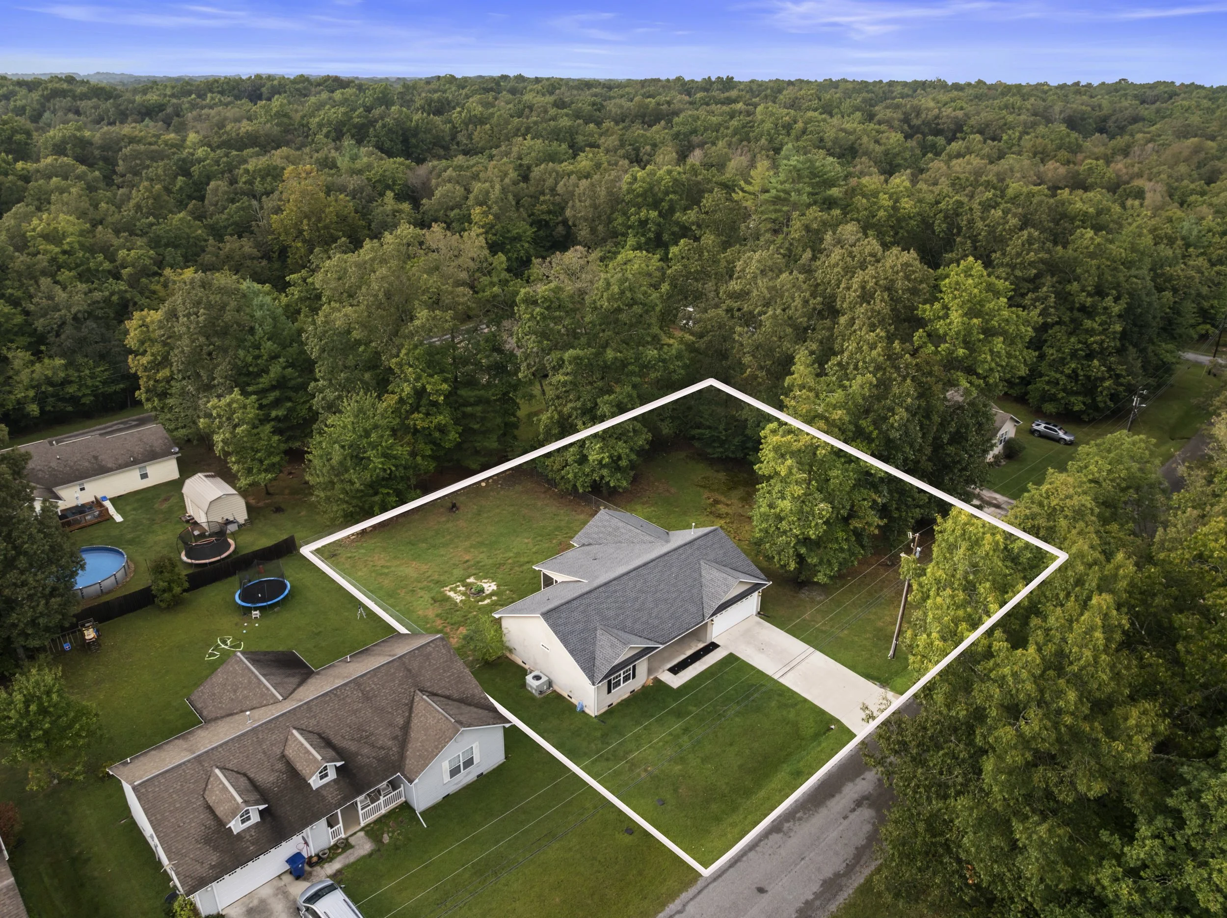 Aerial view of a suburban property with a marked lot boundary, house, lawns, neighboring homes, tree-covered landscape, and blue sky.