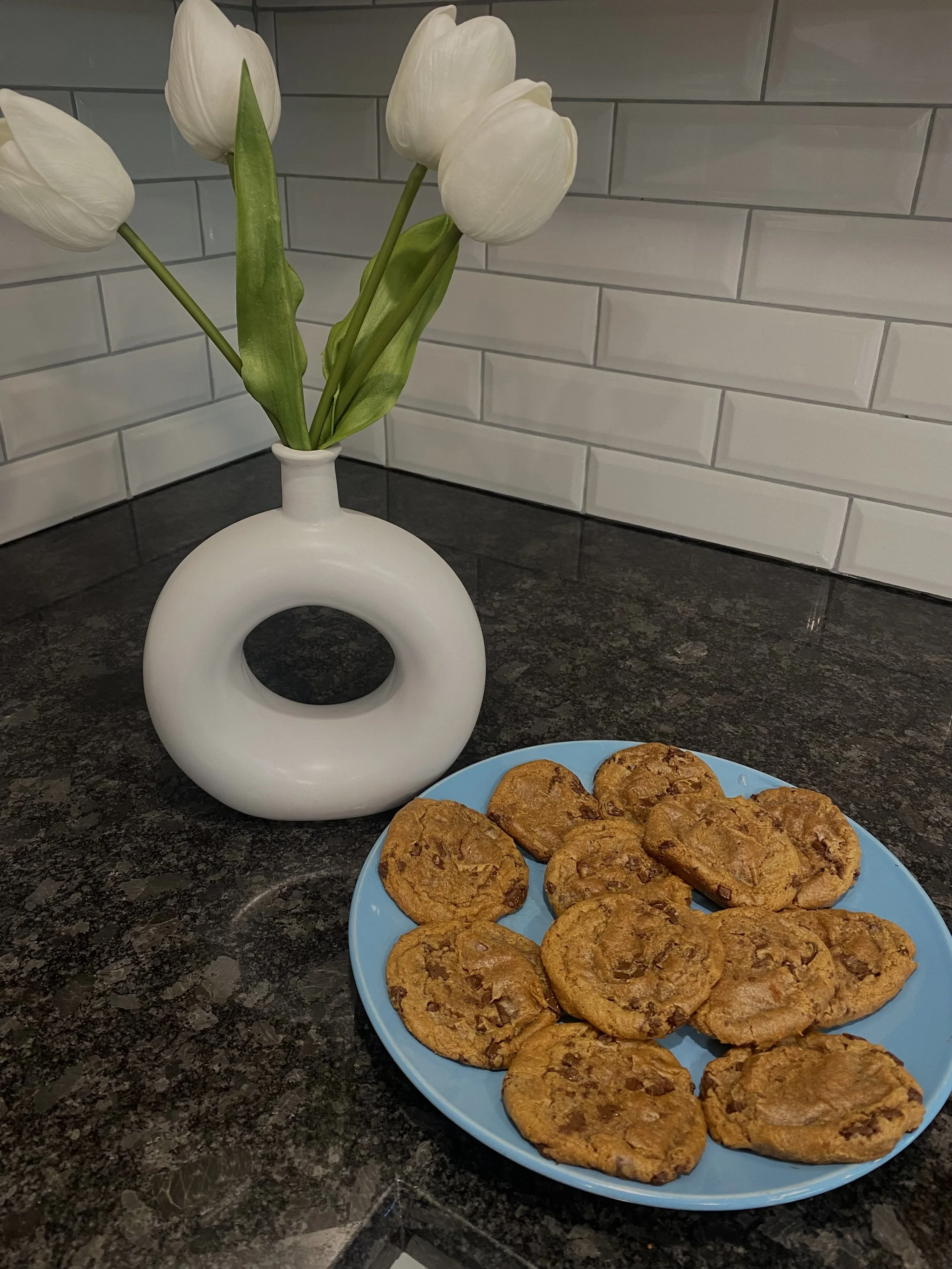 Plate of chocolate chip cookies beside a white vase of tulips on a black granite countertop