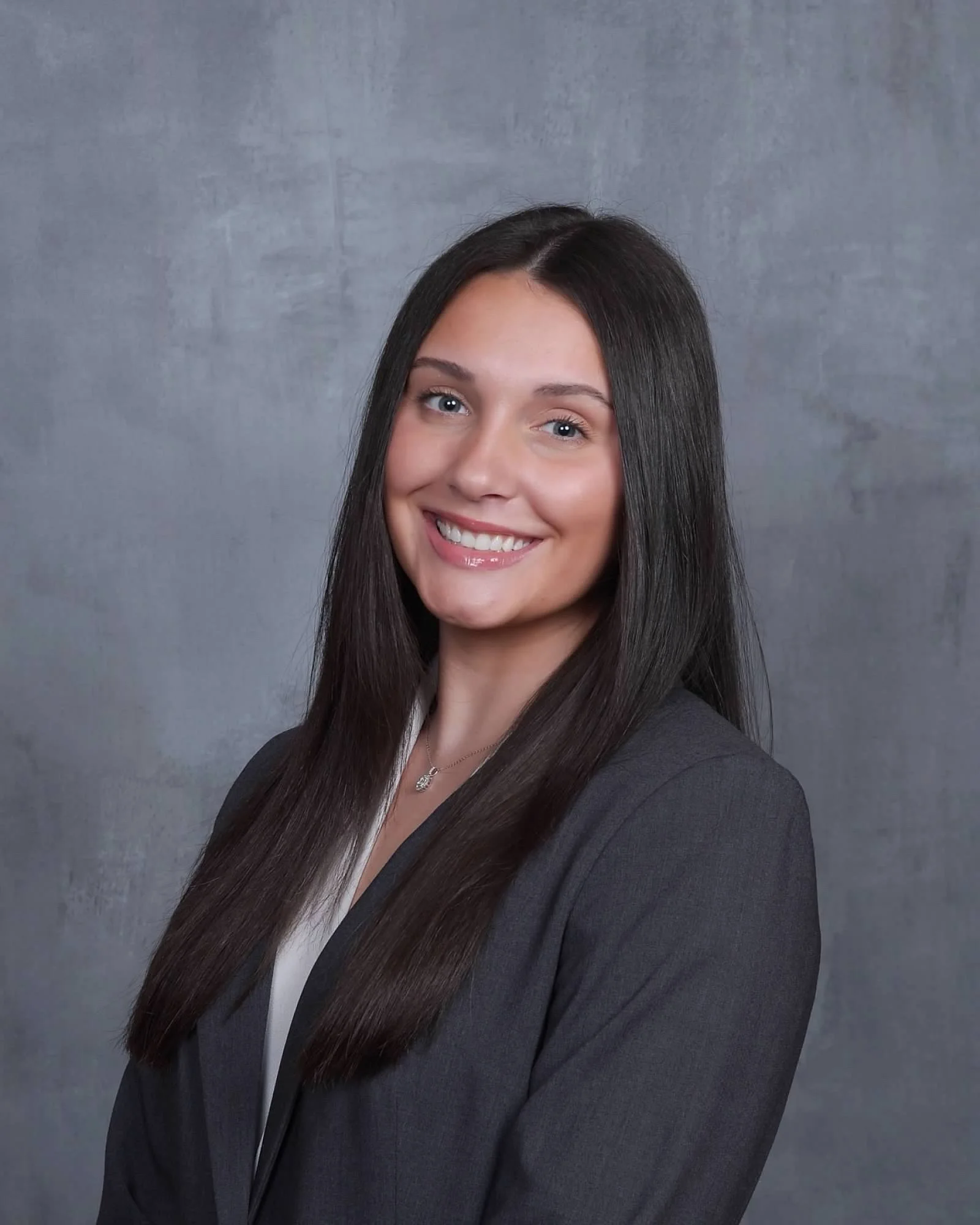 Smiling woman with long dark hair wearing a gray blazer against a gray studio backdrop