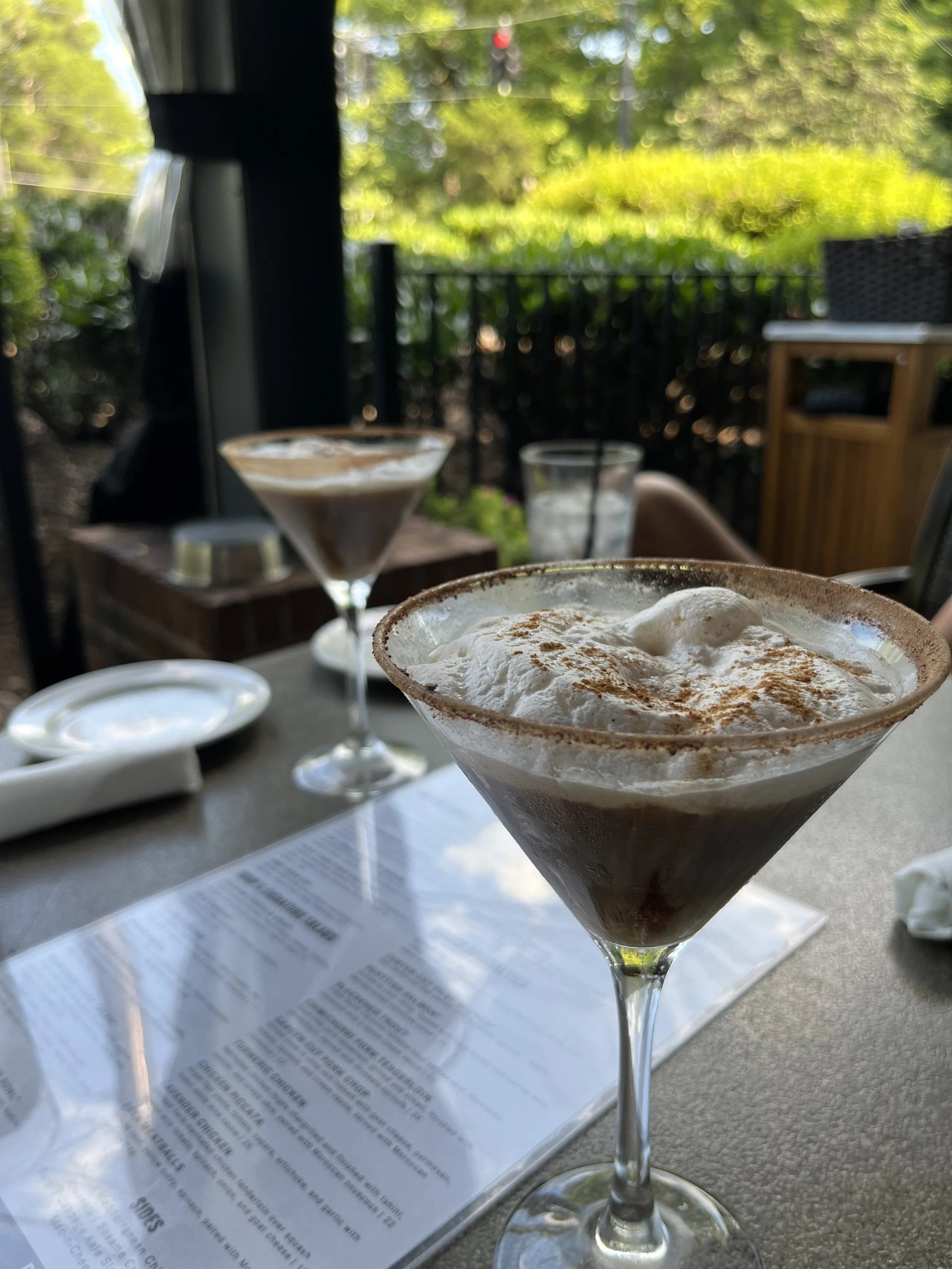 Chocolate dessert cocktail topped with whipped cream and cinnamon on a restaurant table, with a menu and garden patio in the background.