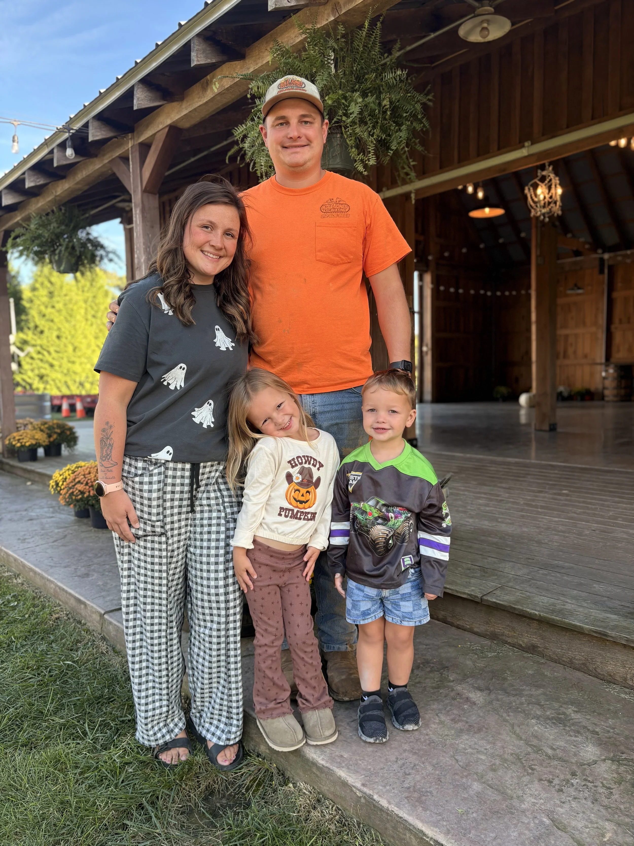 A family of five standing outside a building with wood siding and a covered porch, smiling at the camera. The family includes a woman, a man, and three children, with colorful fall flowers and greenery nearby.