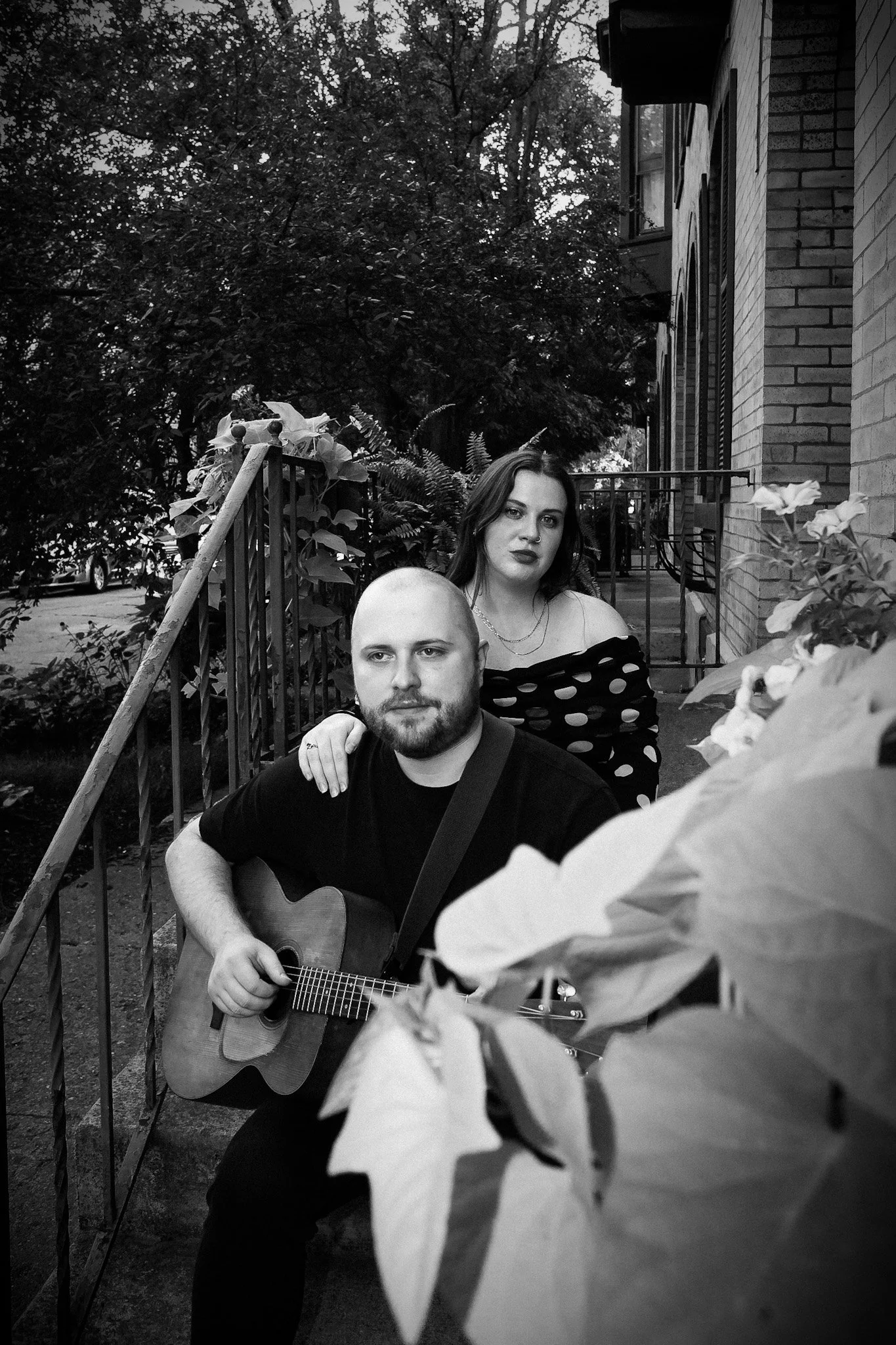 Black-and-white portrait of a man playing acoustic guitar with a woman standing behind him on a leafy residential porch.