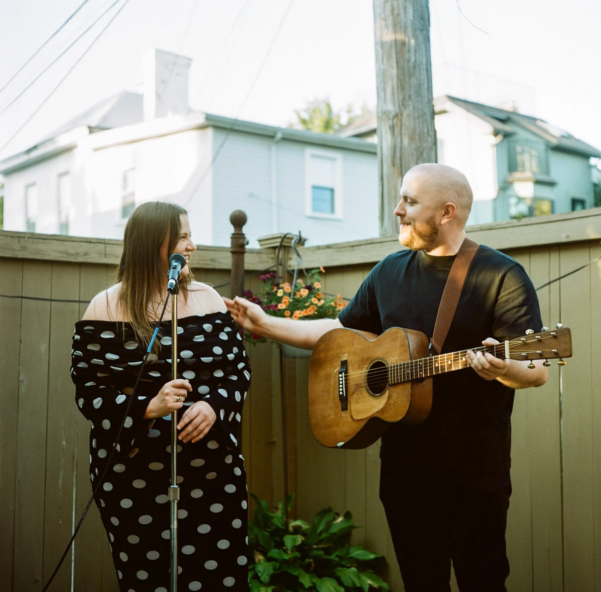 Two musicians perform outdoors, with a woman singing into a microphone and a man playing acoustic guitar.