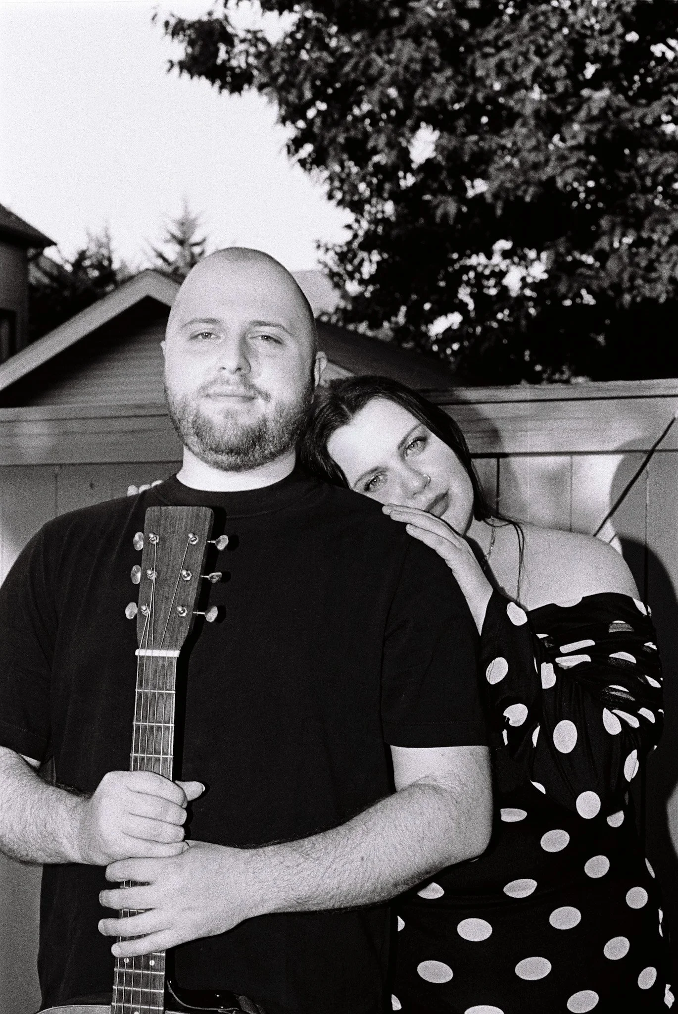 Black-and-white portrait of a couple embracing outdoors, with the man holding an acoustic guitar and the woman wearing a polka-dot dress.
