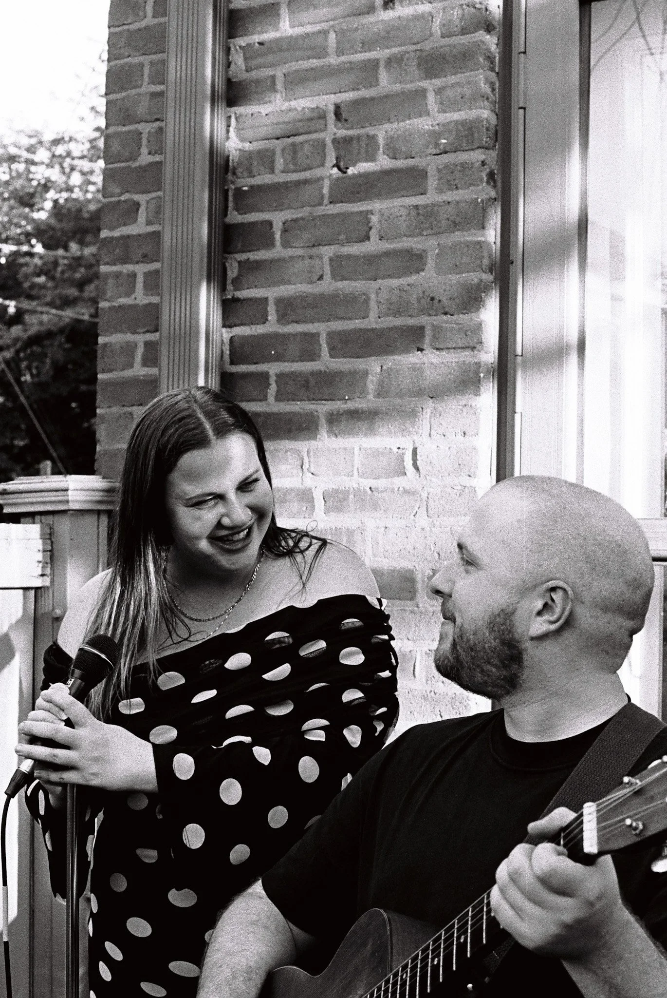 Black-and-white photo of a smiling woman holding a microphone beside a bald man playing acoustic guitar outdoors.