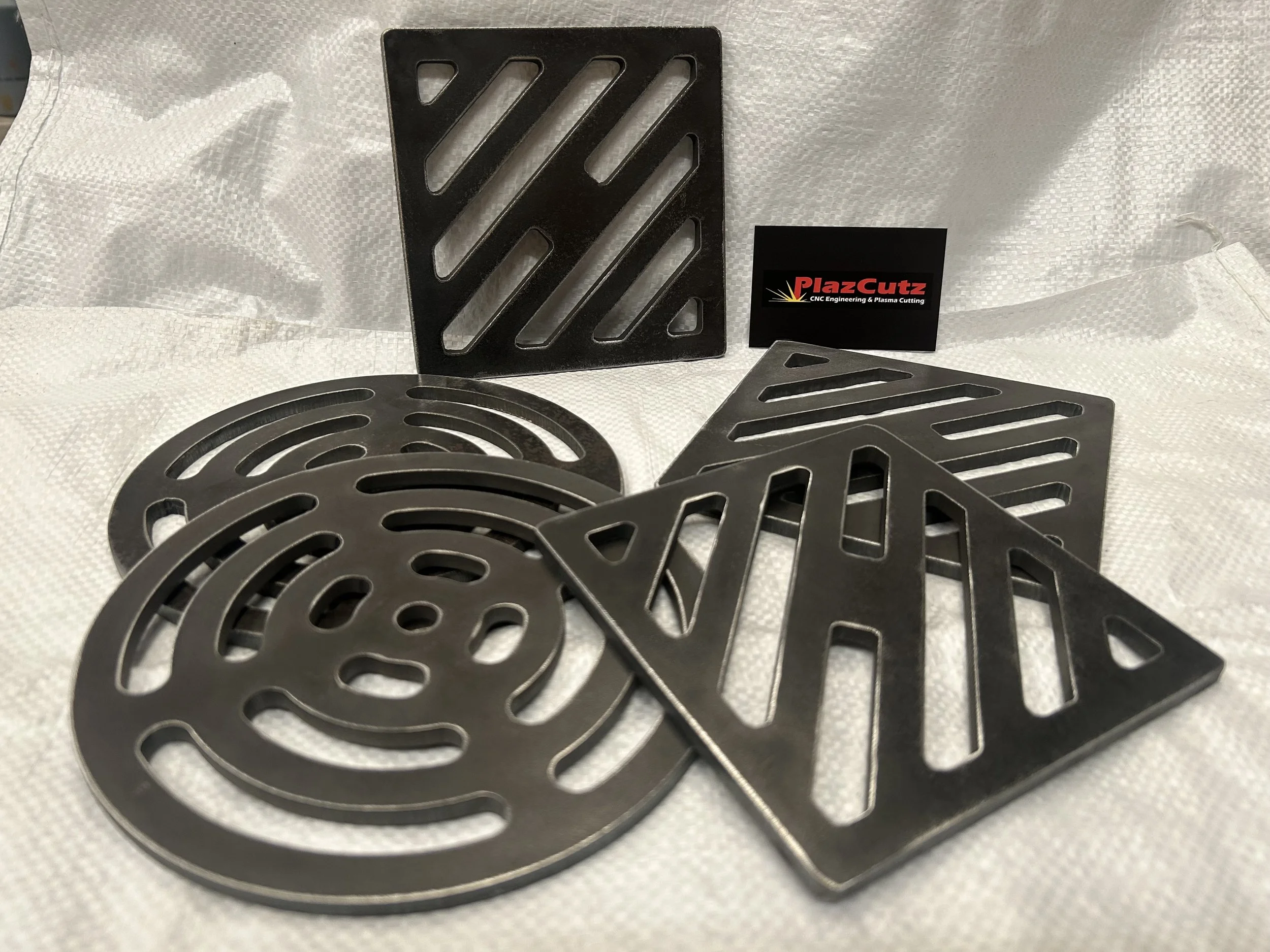 Selection of round and square mild steel drain covers manufactured by PlazCutz, showing CNC plasma cut drainage patterns, bare steel finish and material thickness. Available in 6mm, 8mm and 10mm optio