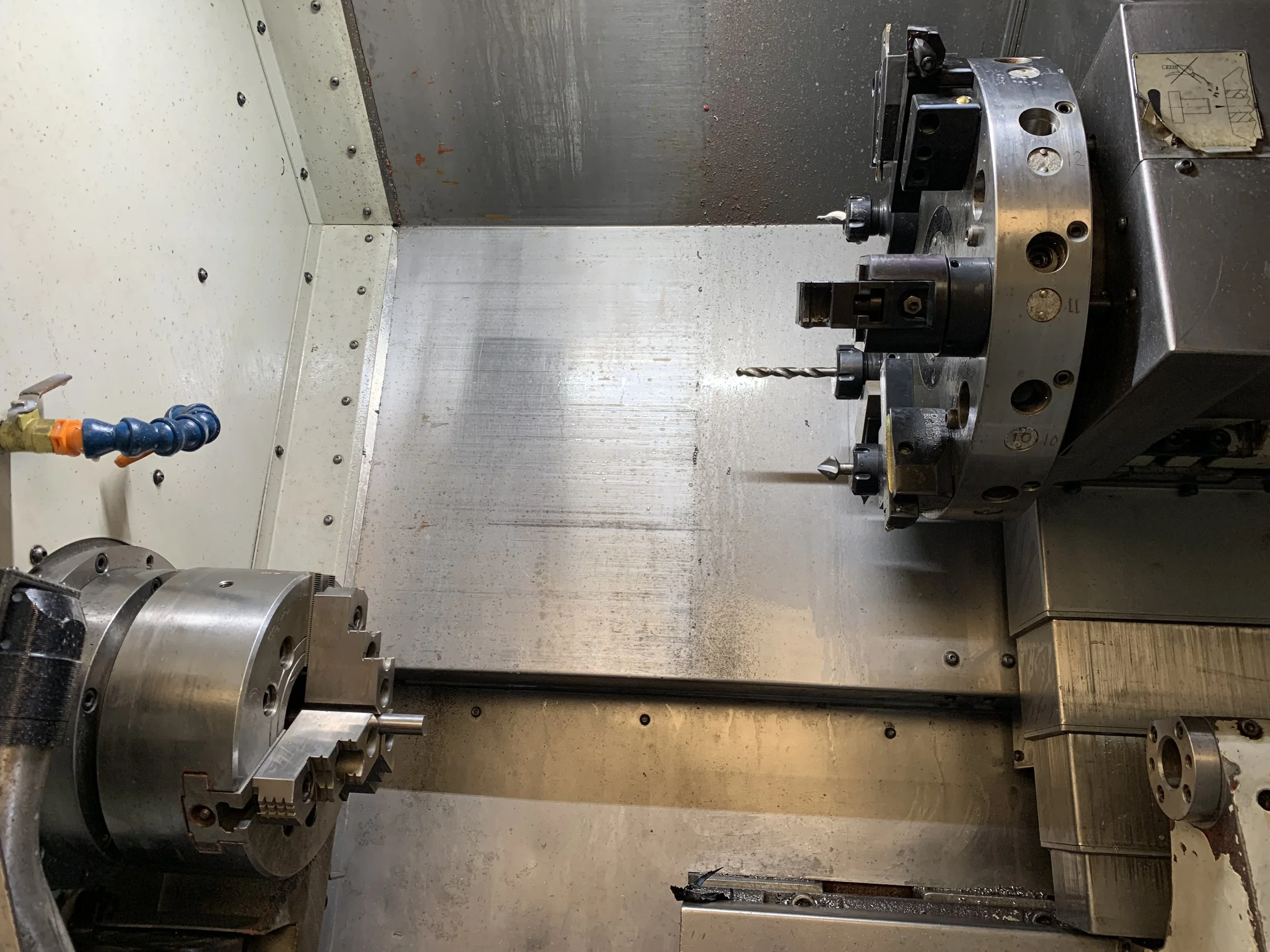 Interior of a CNC lathe with three-jaw chuck, turret-mounted cutting tools, drill bit, and coolant hose.