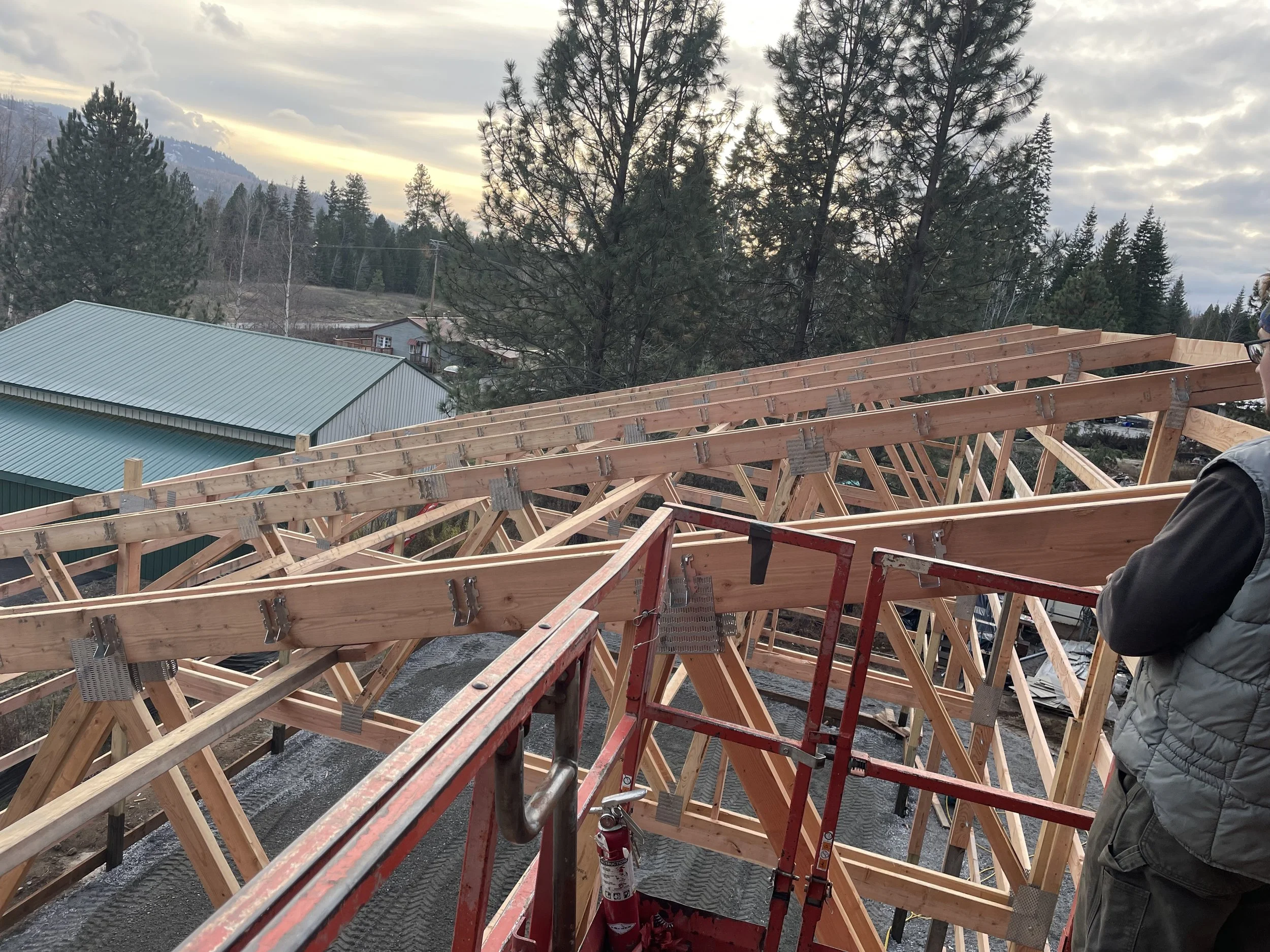 New home roof trusses under construction with workers on a lift, surrounded by pine trees and mountains at sunset