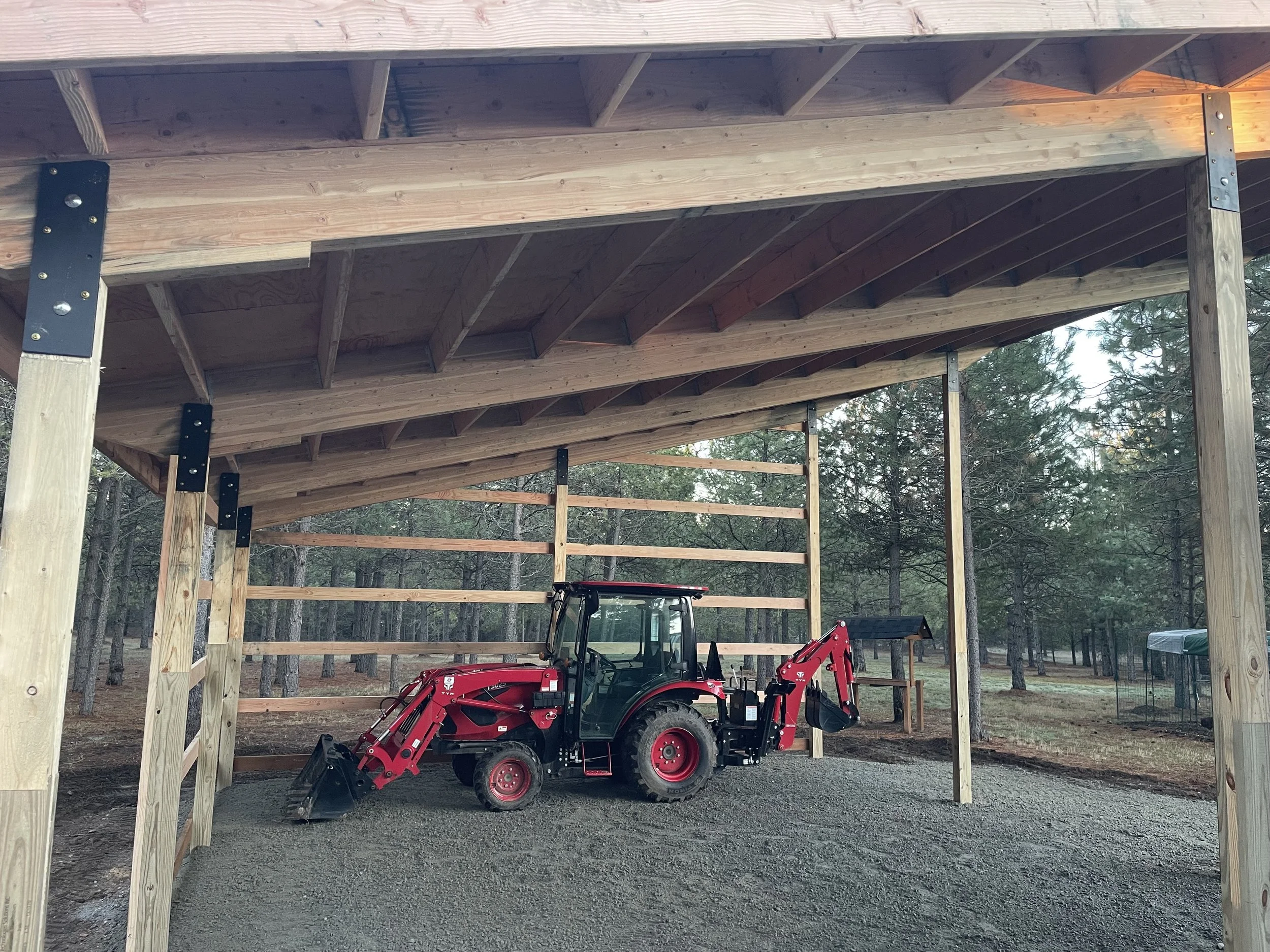 Red compact tractor with front loader and backhoe parked under a wooden shelter in a pine forest