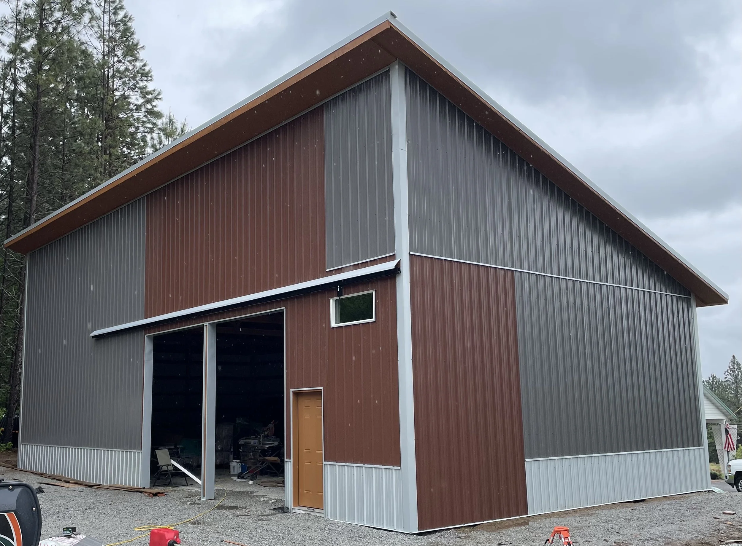 Large red and gray corrugated metal barn or workshop with an open garage door, surrounded by pine trees under a cloudy sky.