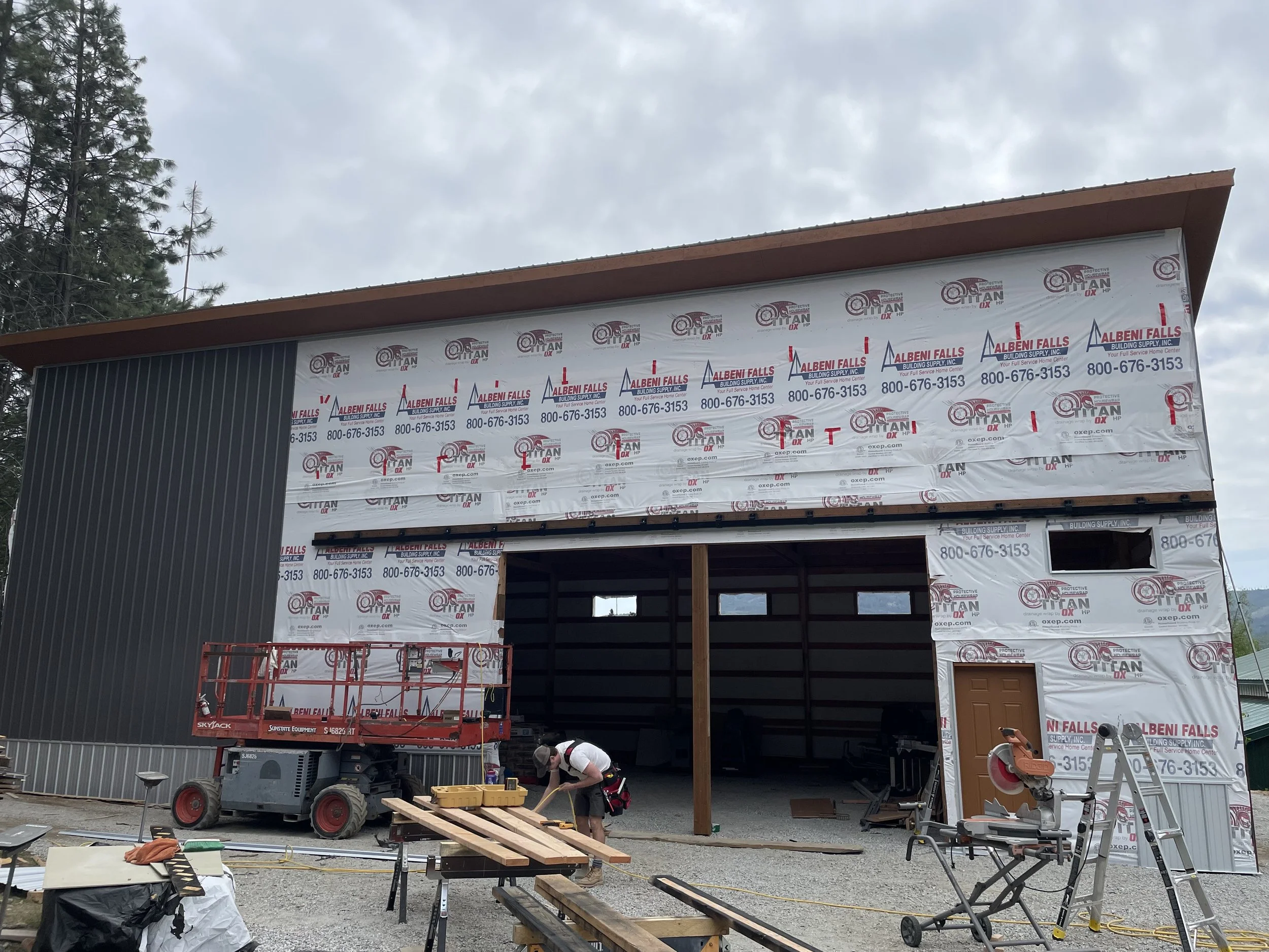 Commercial building under construction with workers, construction equipment, lumber, and weatherproofing wrap labeled Albani Falls Building Supply.