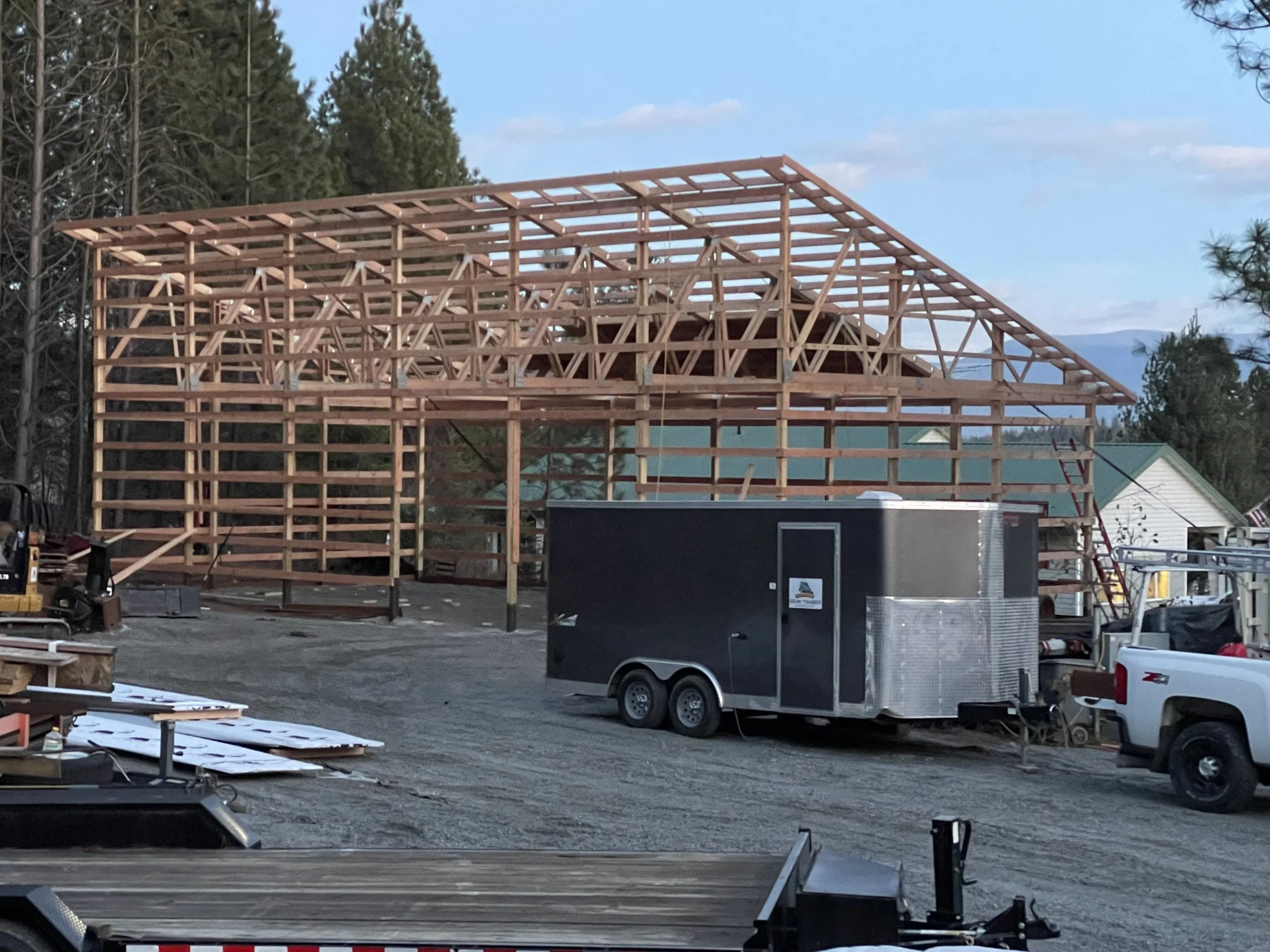 Wooden building under construction with exposed framing, trailer, pickup truck, pine trees, and mountains in the background