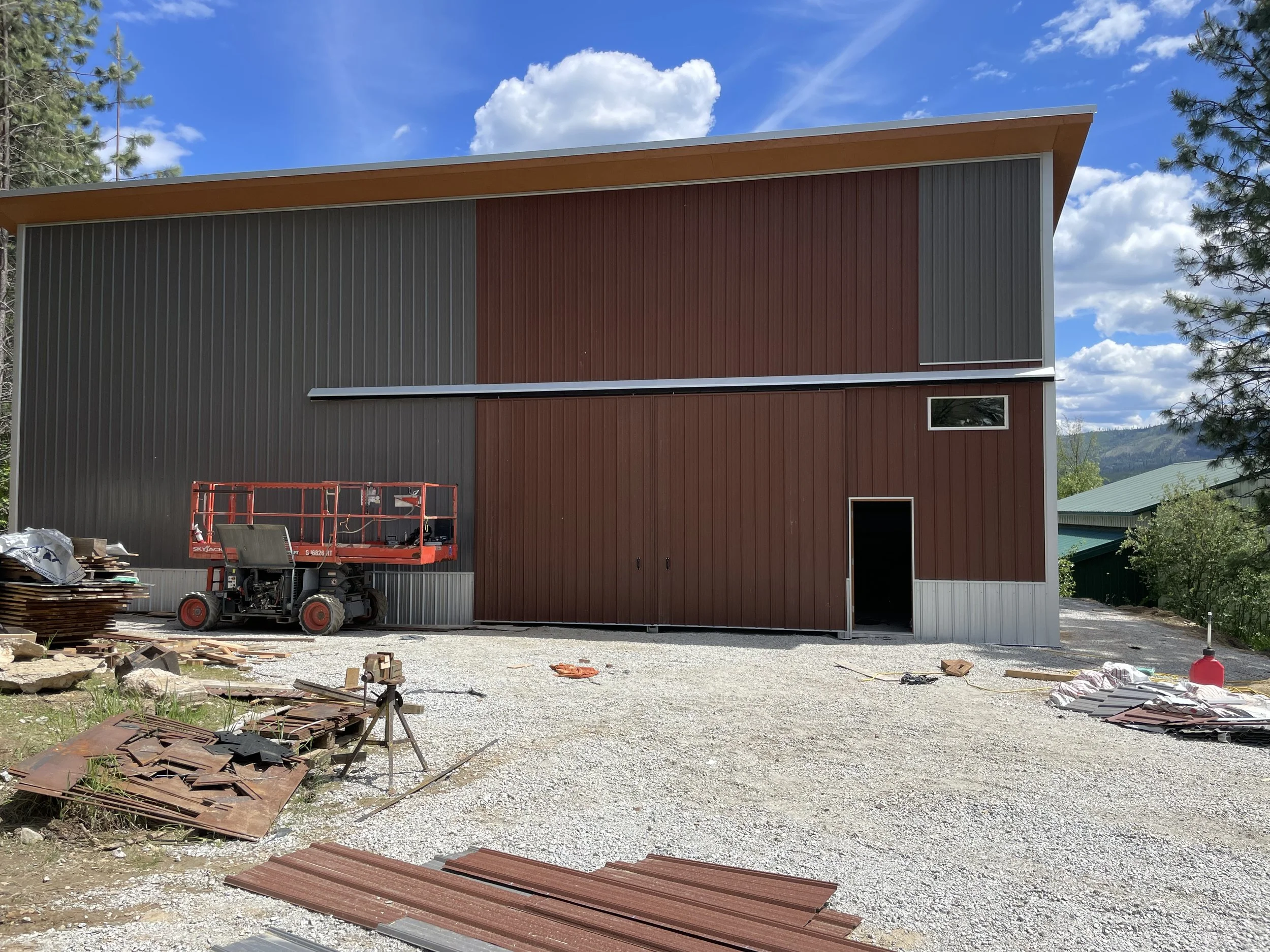 Metal-clad building under construction with a scissor lift, construction materials, pine trees, and mountain views