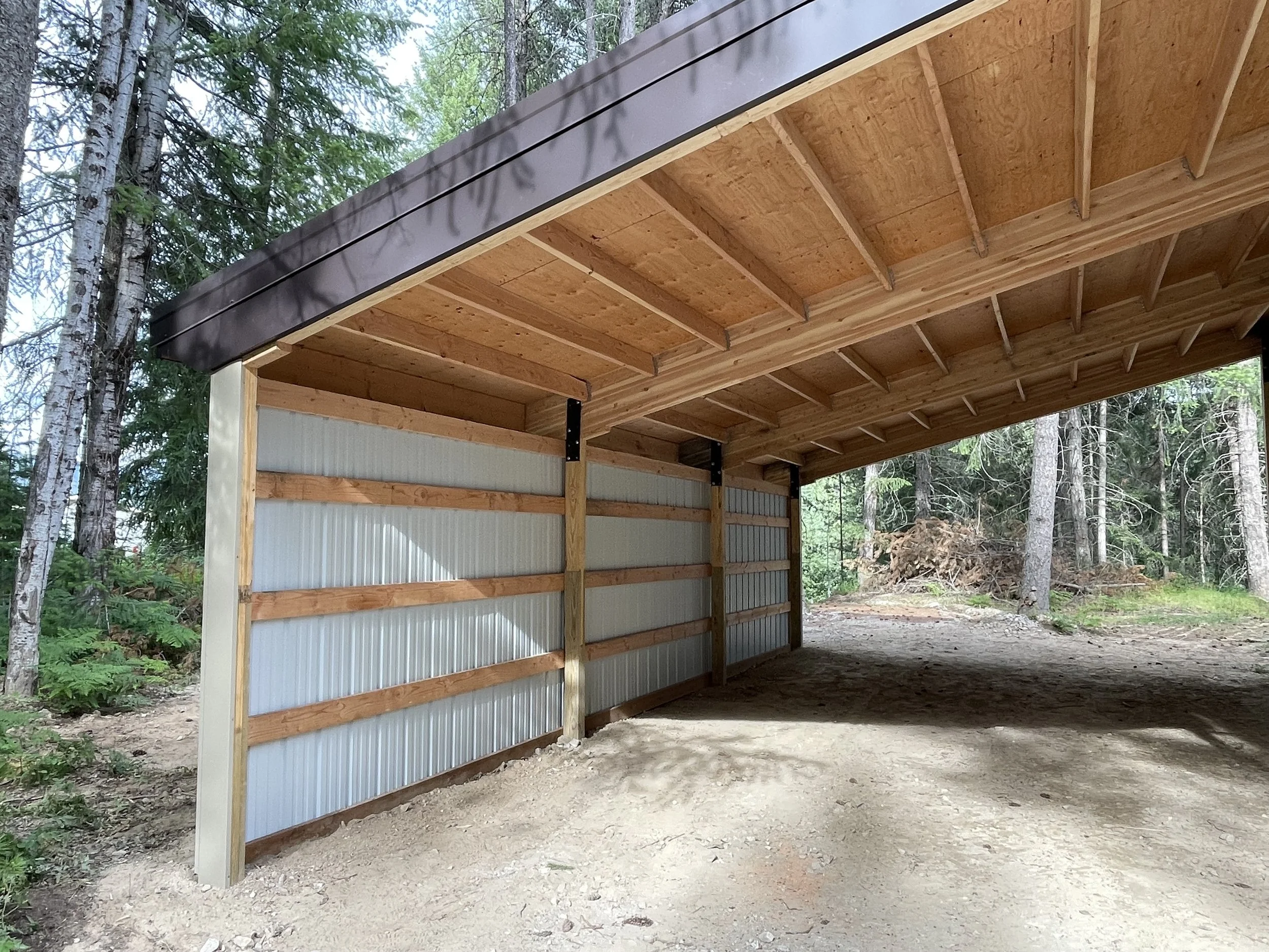 Open-sided wooden carport with corrugated metal walls and roof in a wooded area