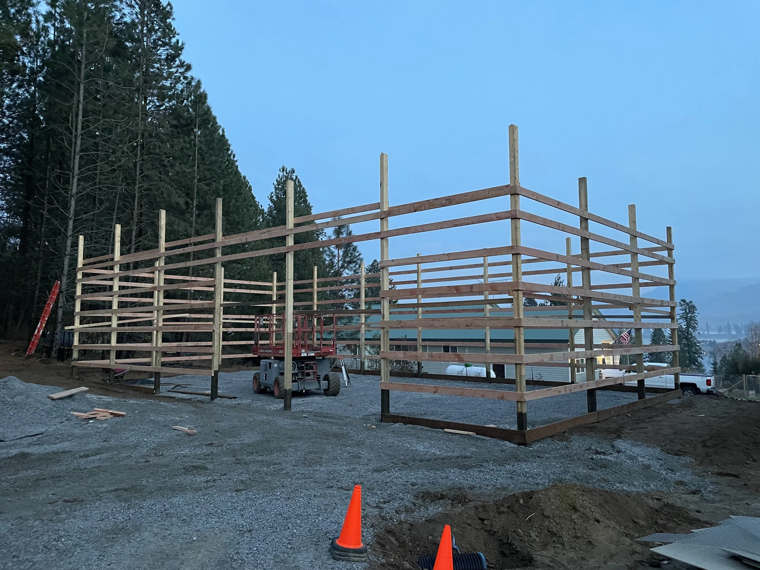 Timber-frame building under construction beside pine trees, with construction equipment, orange traffic cones, and a mountain view