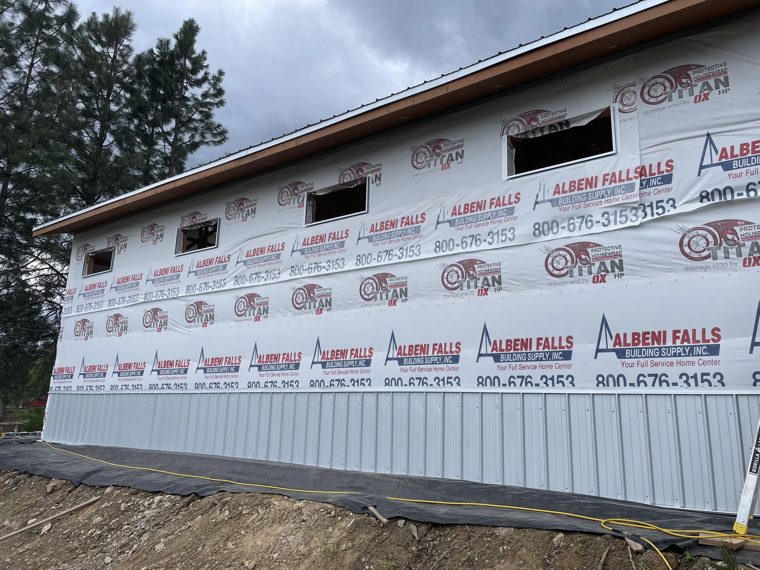 House under construction wrapped in Titan drainage housewrap with multiple windows, metal siding, and Albeni Falls Building Supply branding