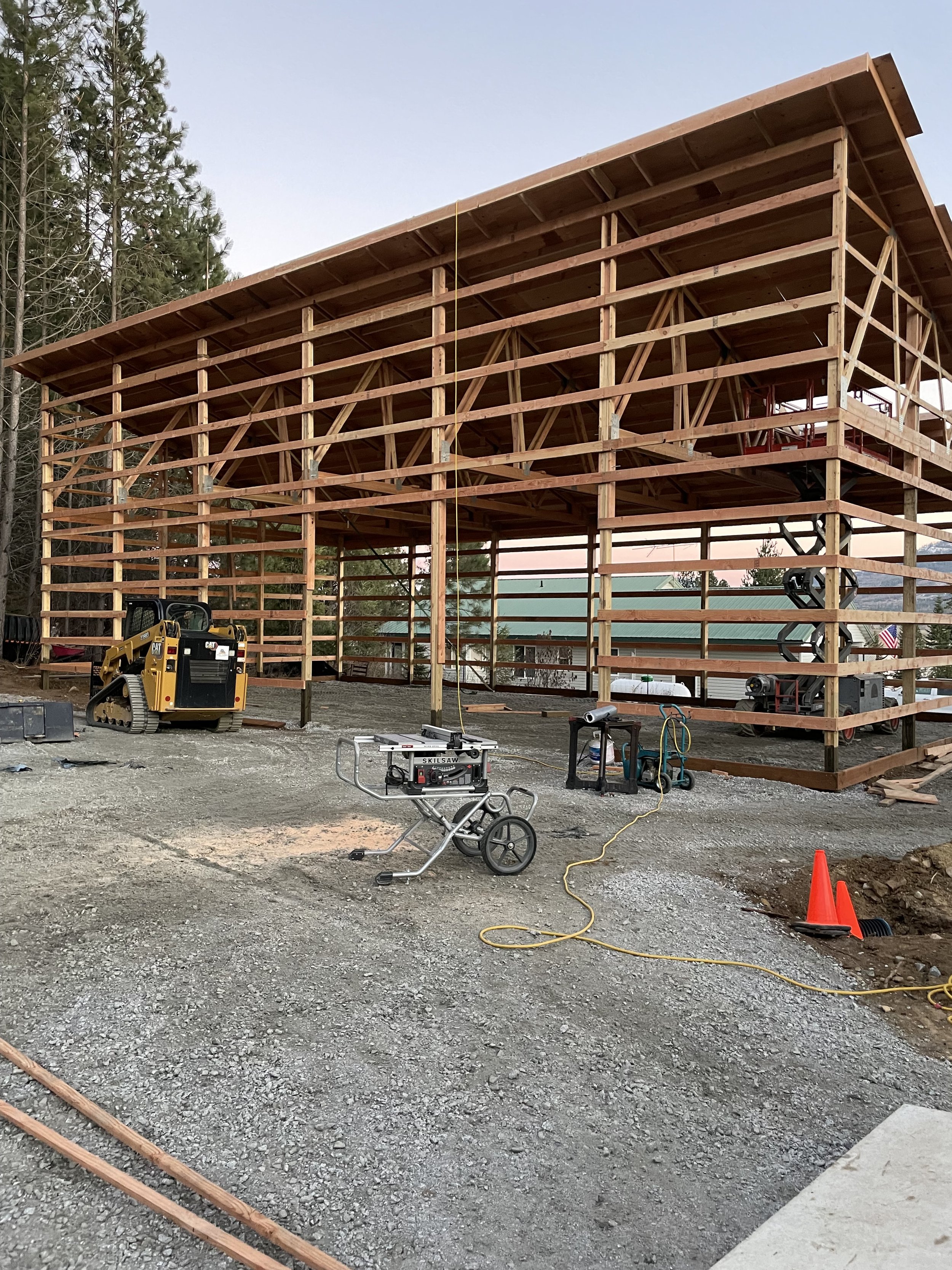 Wooden building under construction with heavy equipment, a portable table saw, tools, and orange safety cones on a gravel site.
