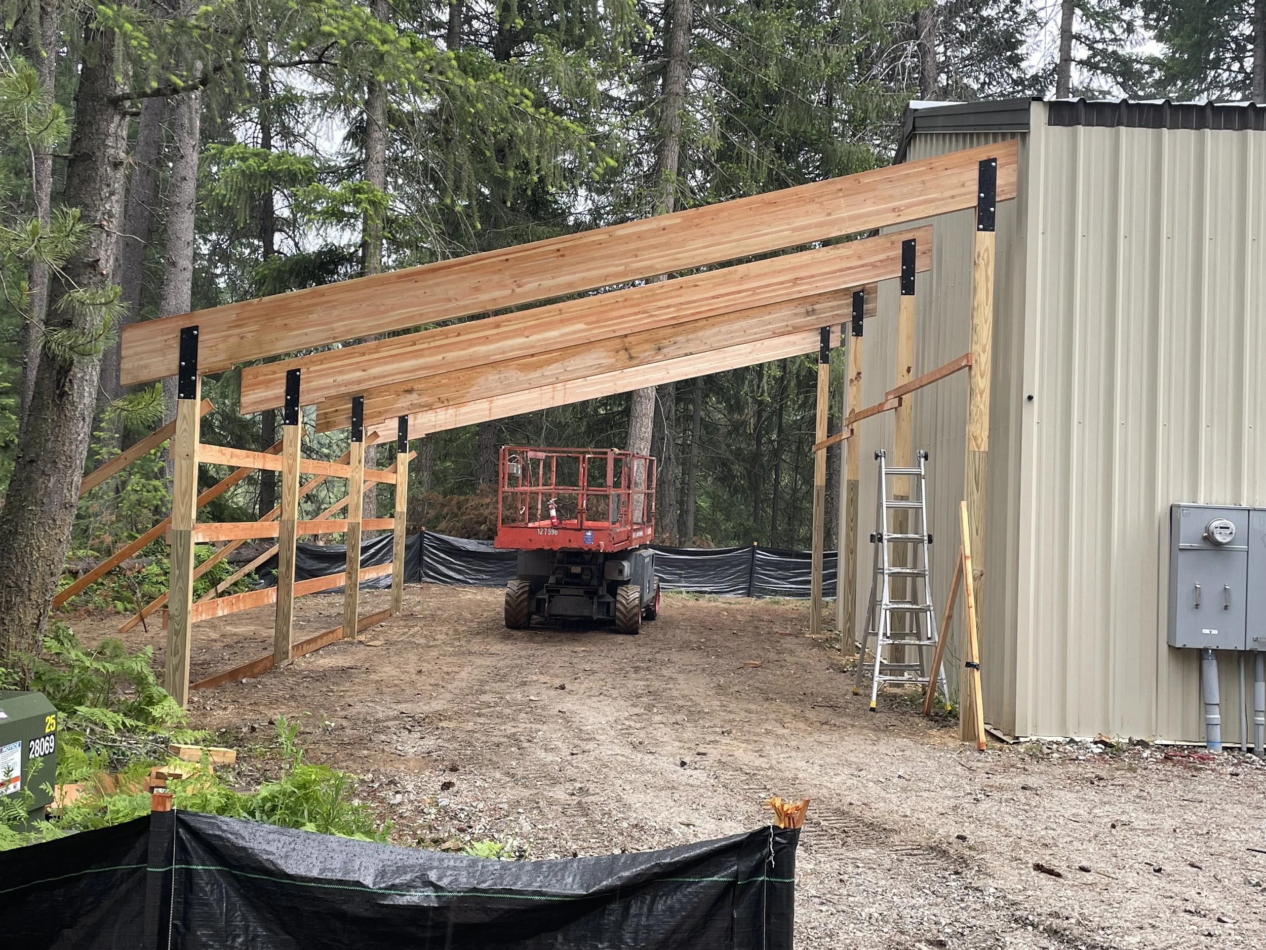 Wooden covered walkway under construction beside a metal building in a wooded area, with a lift and ladder onsite