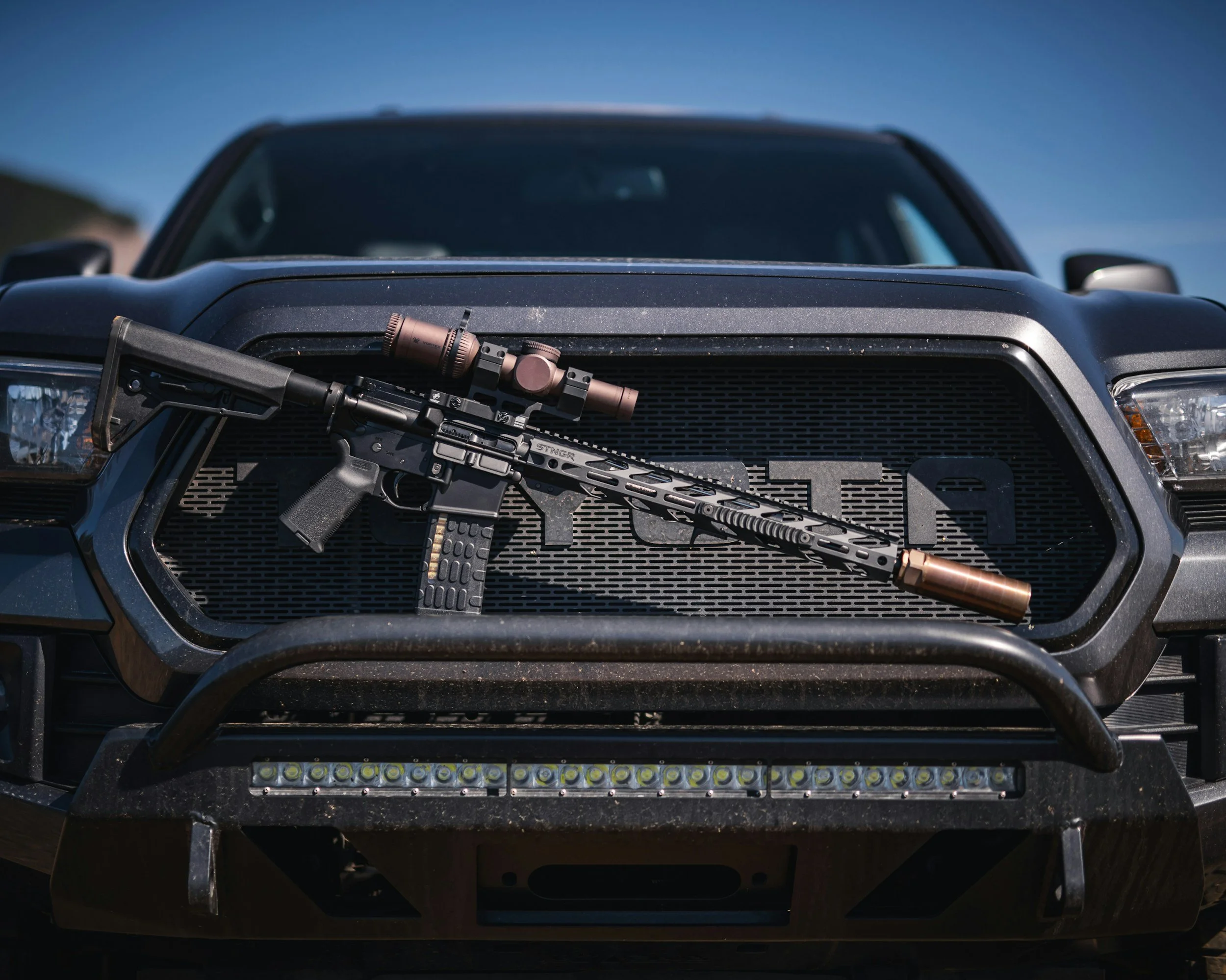 Modern sporting rifle with scope and suppressor displayed on the front grille of a black off-road vehicle