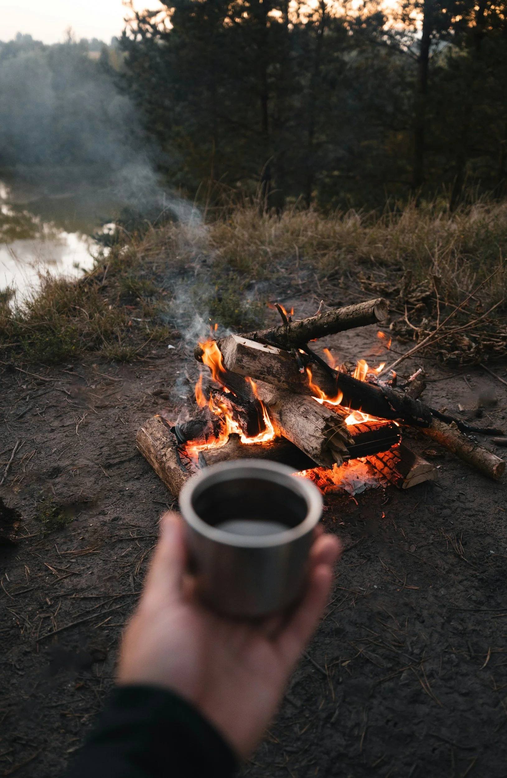 A person holding a coffee cup near a campfire in a wooded area during dusk.
