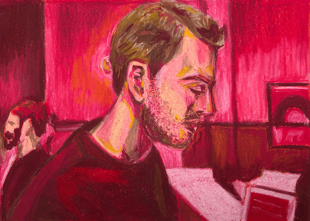Retrato de um homem jovem com barba e cabelo castanho claro, olhando para a tela de um computador, com fundo de parede rosa e uma outra pessoa ao fundo.