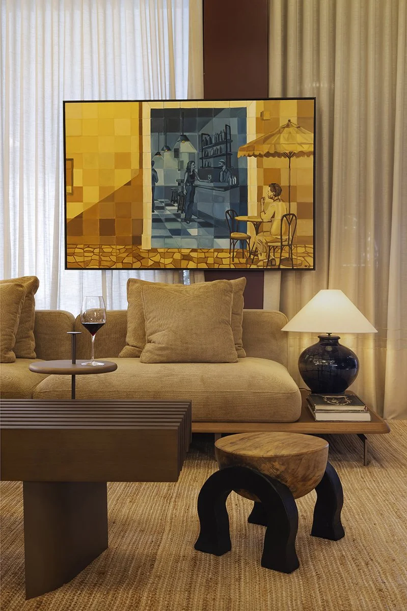 Sala de estar com sofá bege, mesa de vinho, lâmpada de mesa com base escura, painel de TV com quadro de um café ao ar livre, cortinas brancas e parede marrom ao fundo.