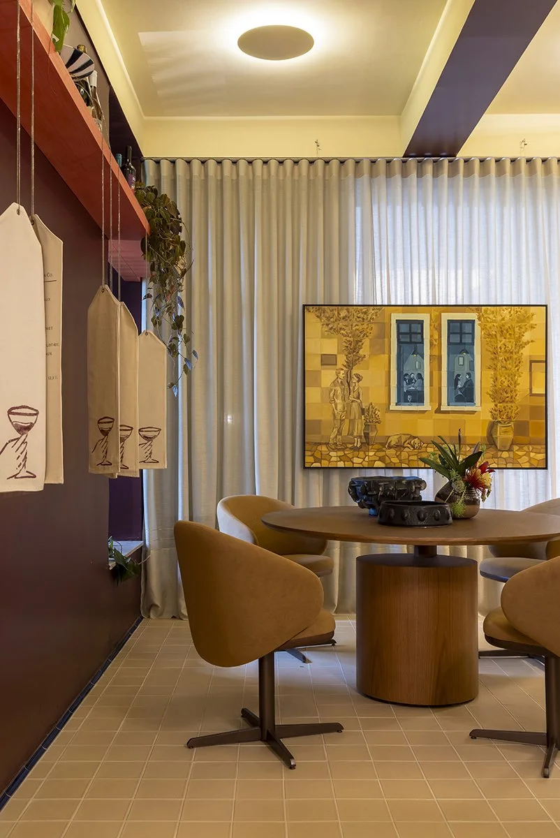 Ambiente de sala de jantar com mesa de madeira e quatro cadeiras estofadas em tom bege. Ao fundo, uma pintura com elementos arquitetônicos e plantas. Cortinas brancas e luminária no teto oferecem iluminação suave.