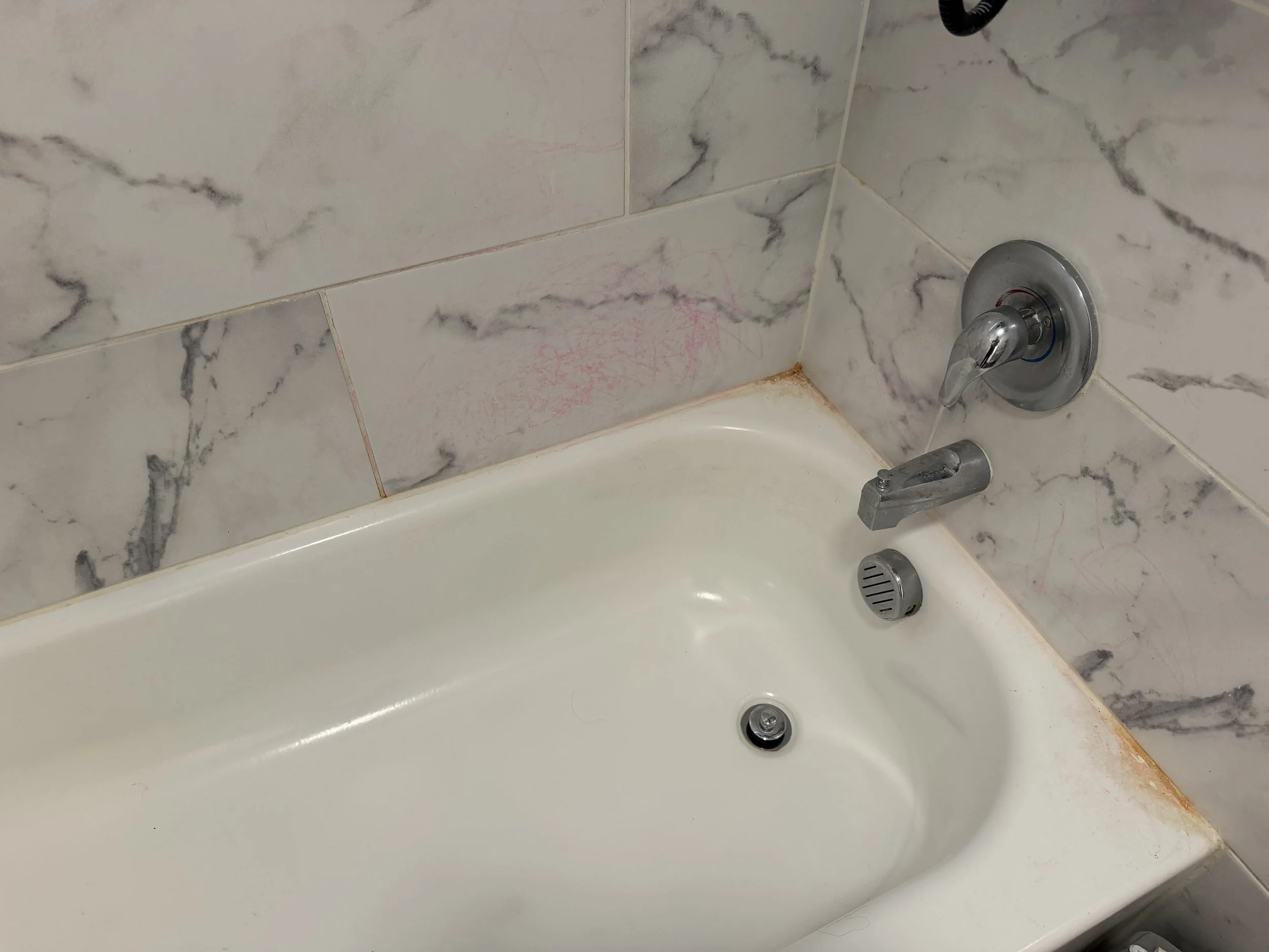 White bathtub with chrome faucet and drain, surrounded by marble-patterned tile and visible pink stains and rust along the caulked edges.