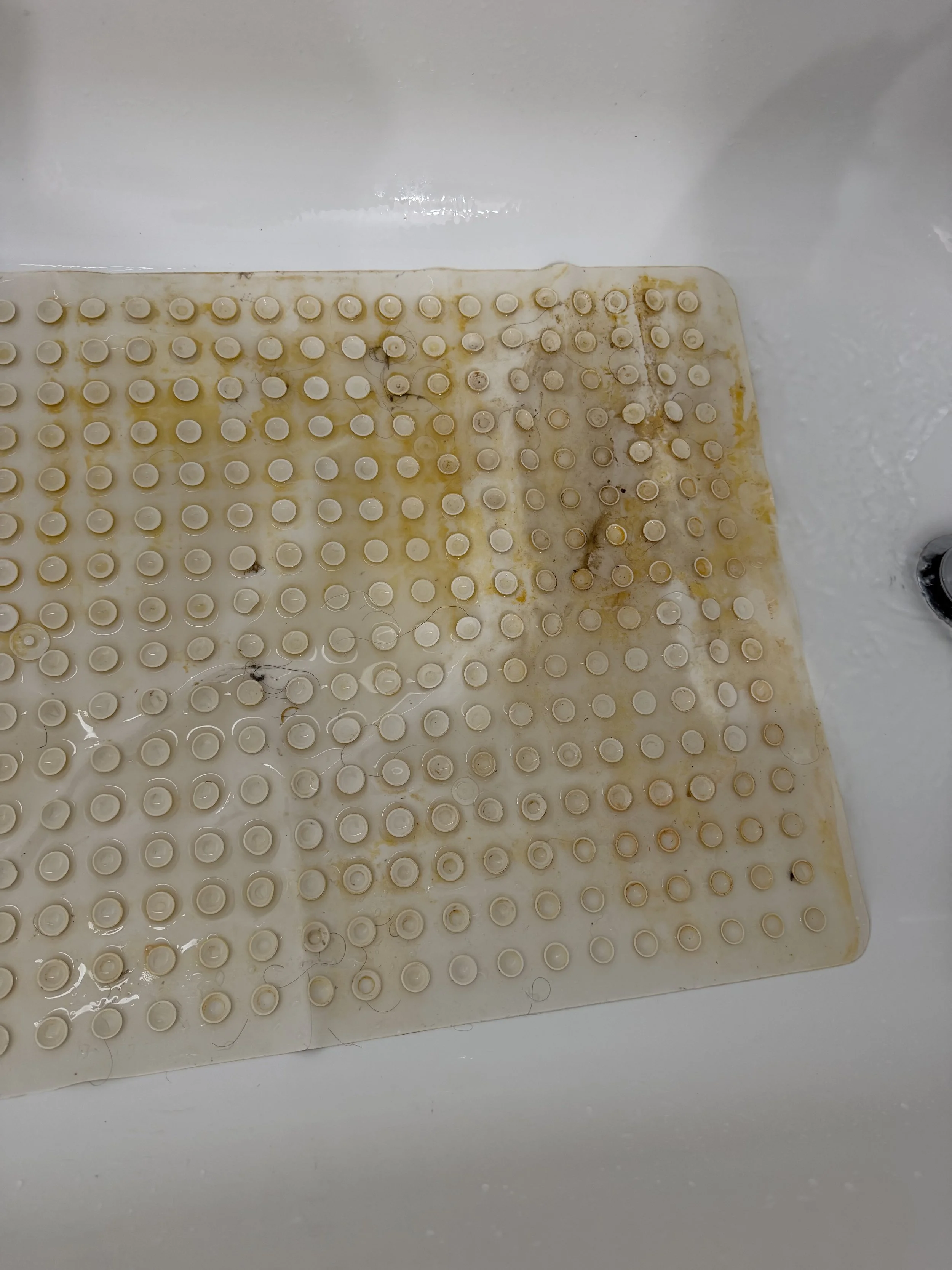Dirty, stained white bathtub mat with rows of suction cups and scattered hair.
