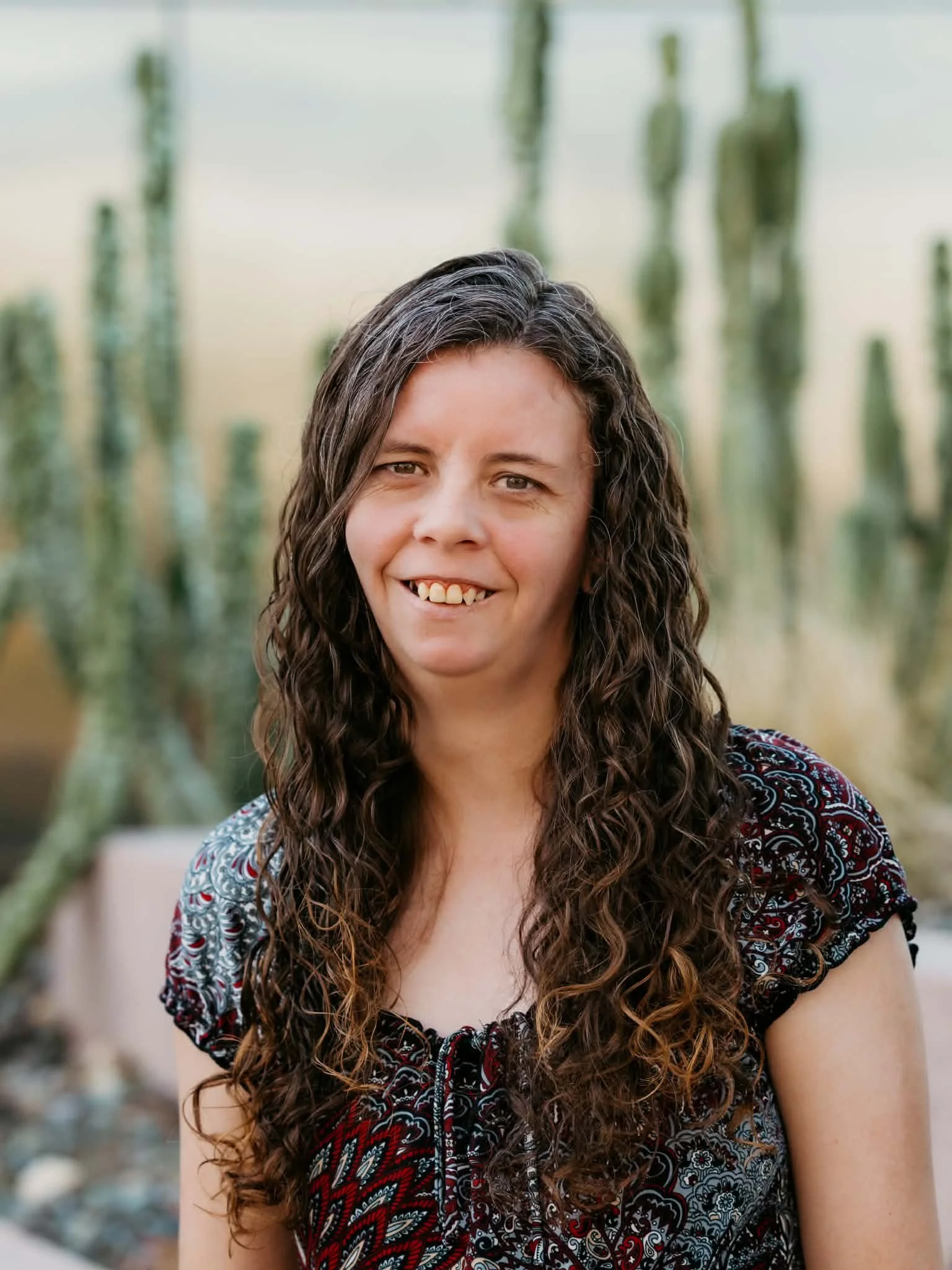 Smiling woman with long curly brown hair wearing a patterned top, photographed outdoors with blurred cacti in the background