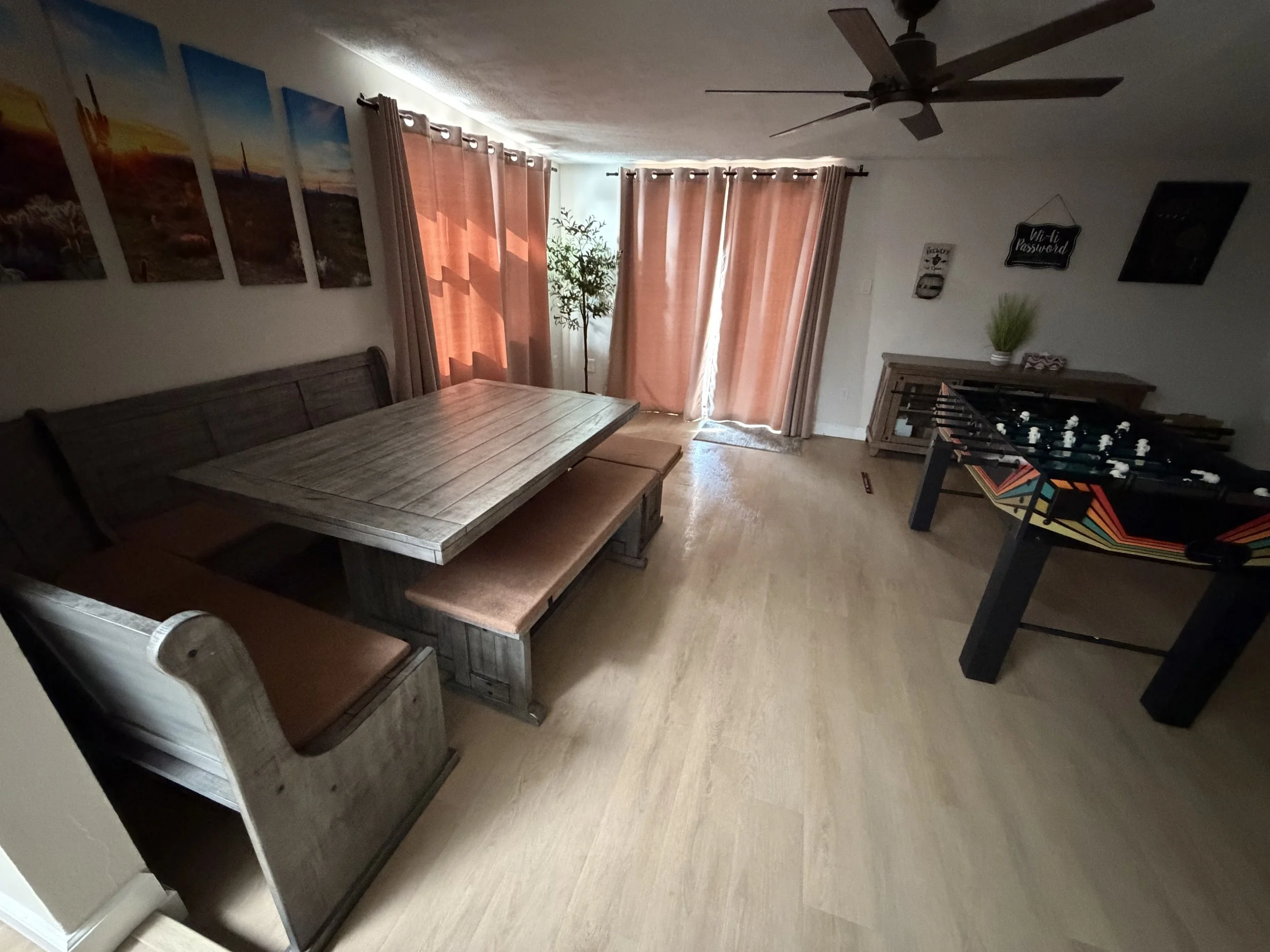 Bright dining and game room with a rustic wood table and bench seating, foosball table, ceiling fan, framed wall art, and pink curtains.