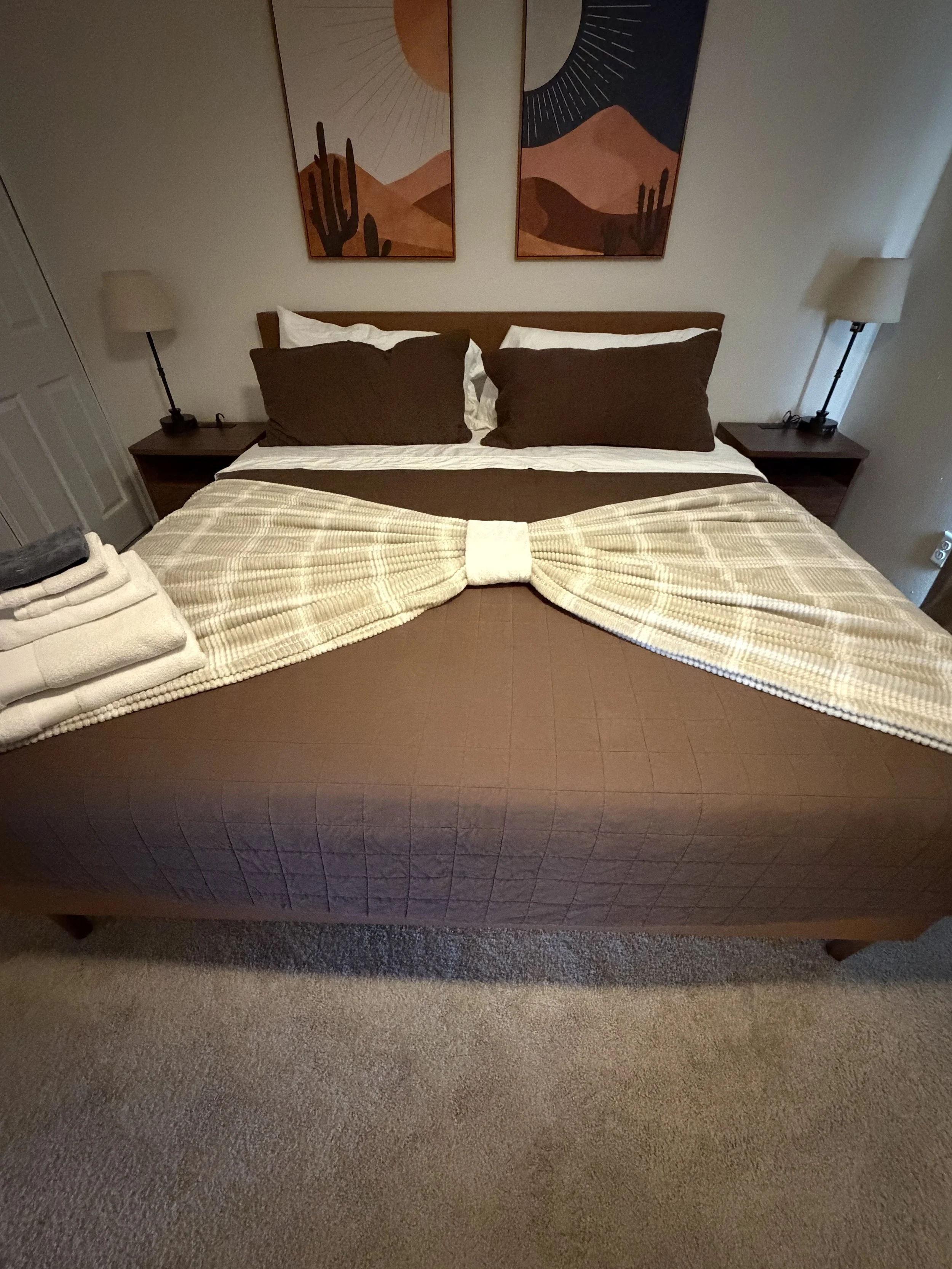 Cozy bedroom with a neatly made brown bed, white textured throw, matching nightstands and lamps, and desert landscape artwork above the headboard.