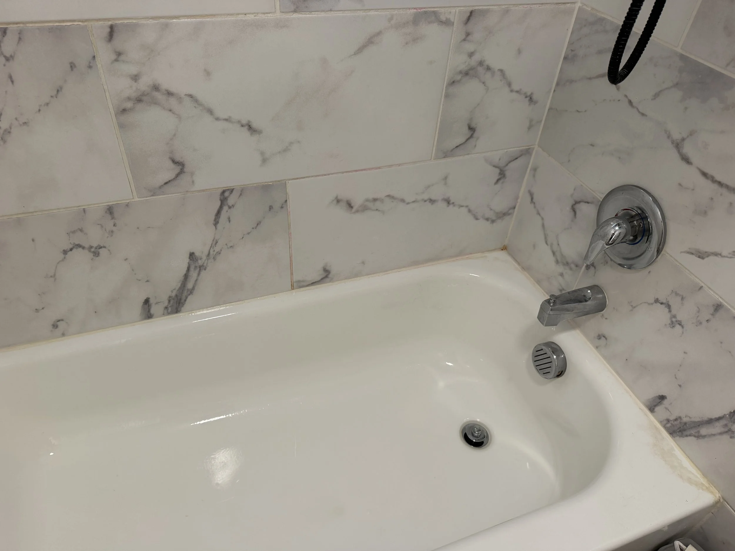 White bathtub with chrome faucet and drain set against marble-patterned tile walls.