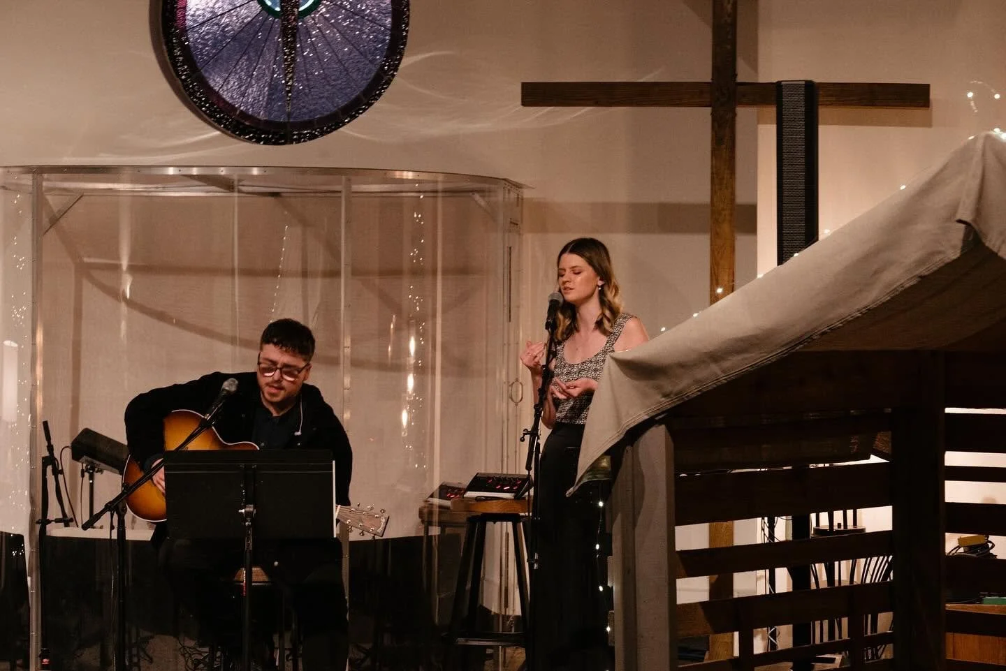 Musicians performing inside a church, with a guitarist, female singer, large cross, and stained-glass window