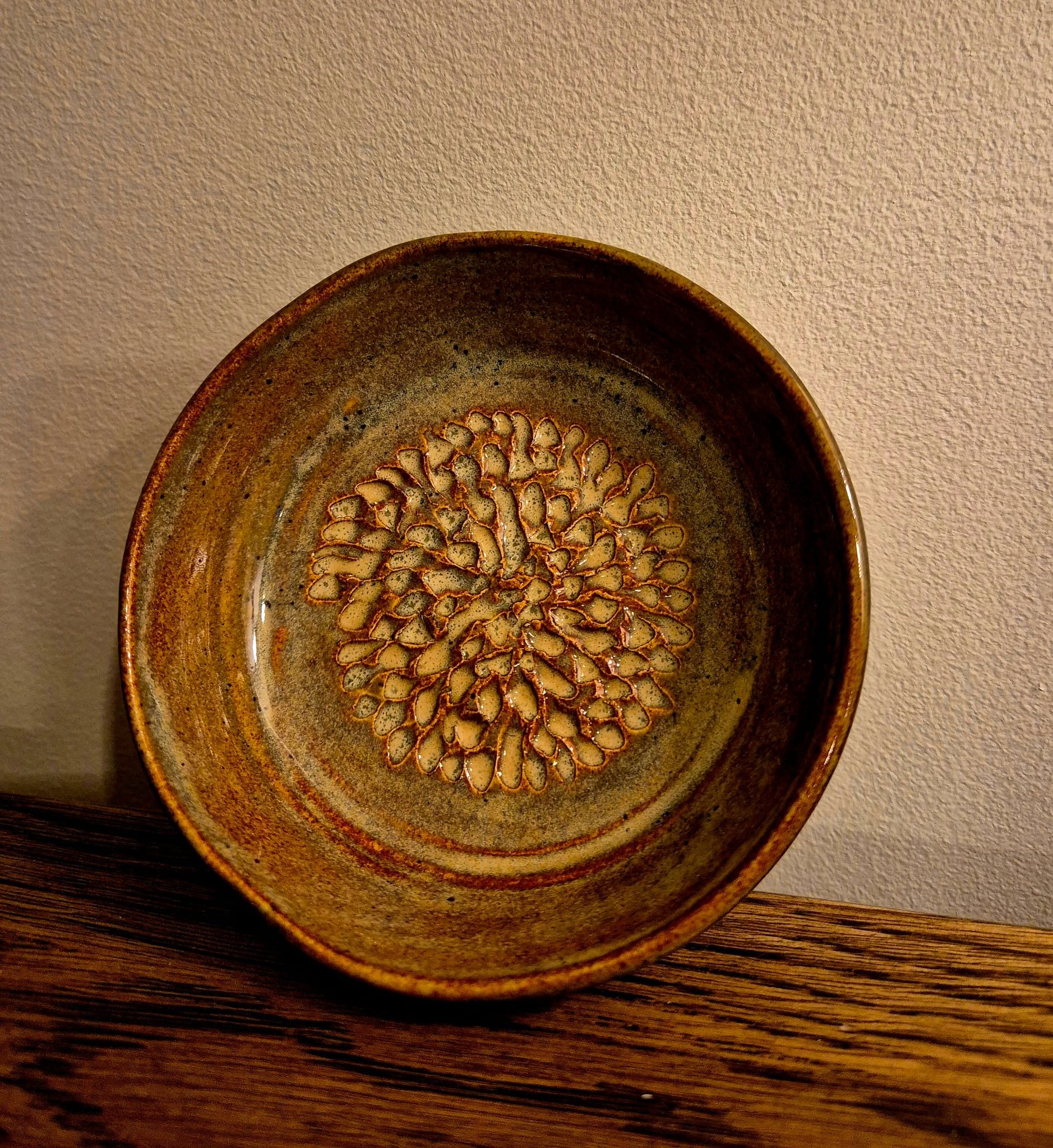 Large rustic brown glazed ceramic bowl with a floral pattern, displayed on a wooden table against a textured wall.