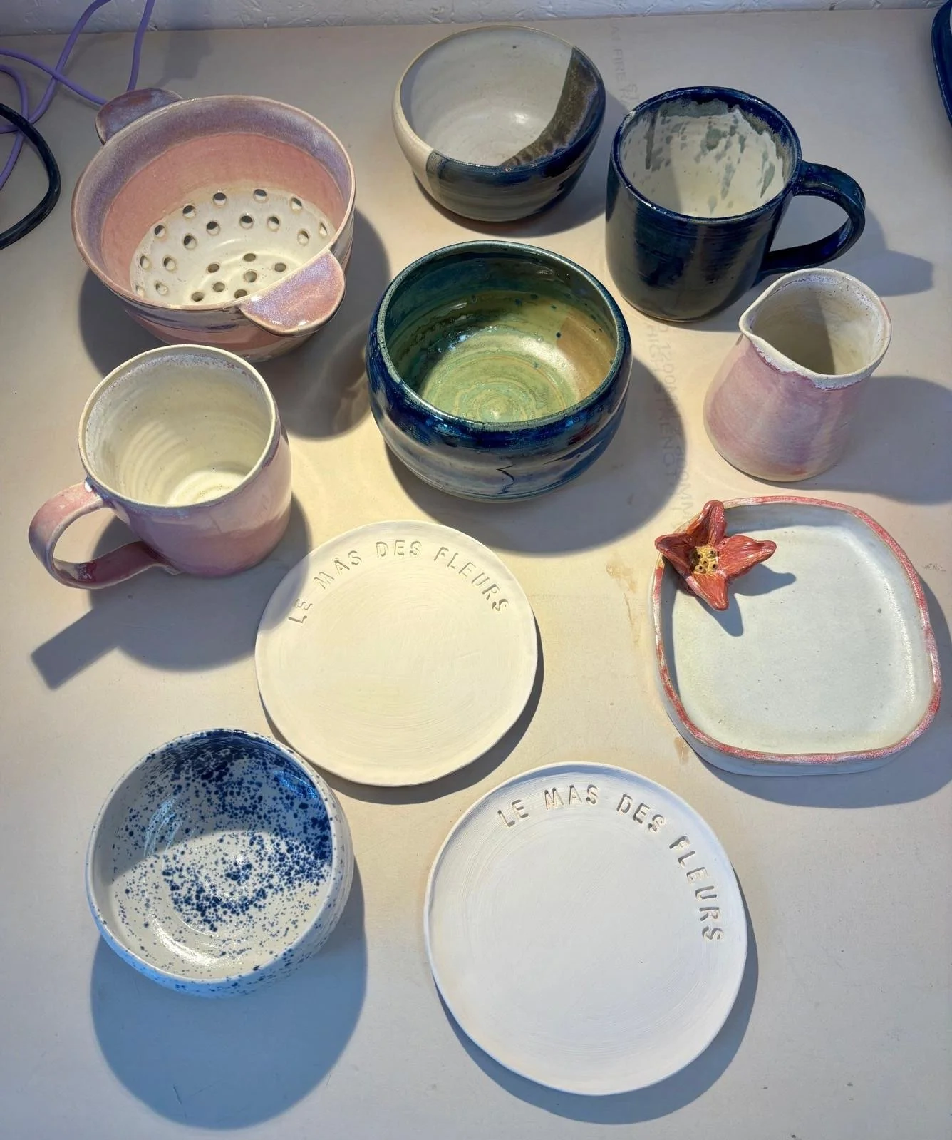 Assortment of handmade ceramic pottery, including colorful bowls, mugs, a colander, pitchers, and decorative plates arranged on a sunlit table