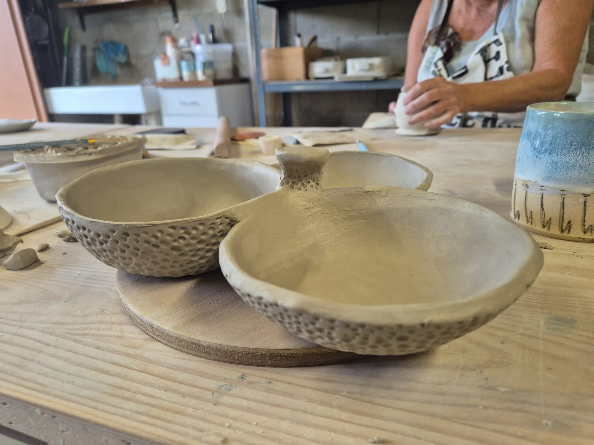 Handcrafted ceramic bowls and a blue-and-white mug on a pottery studio worktable, with an artist shaping clay in the background
