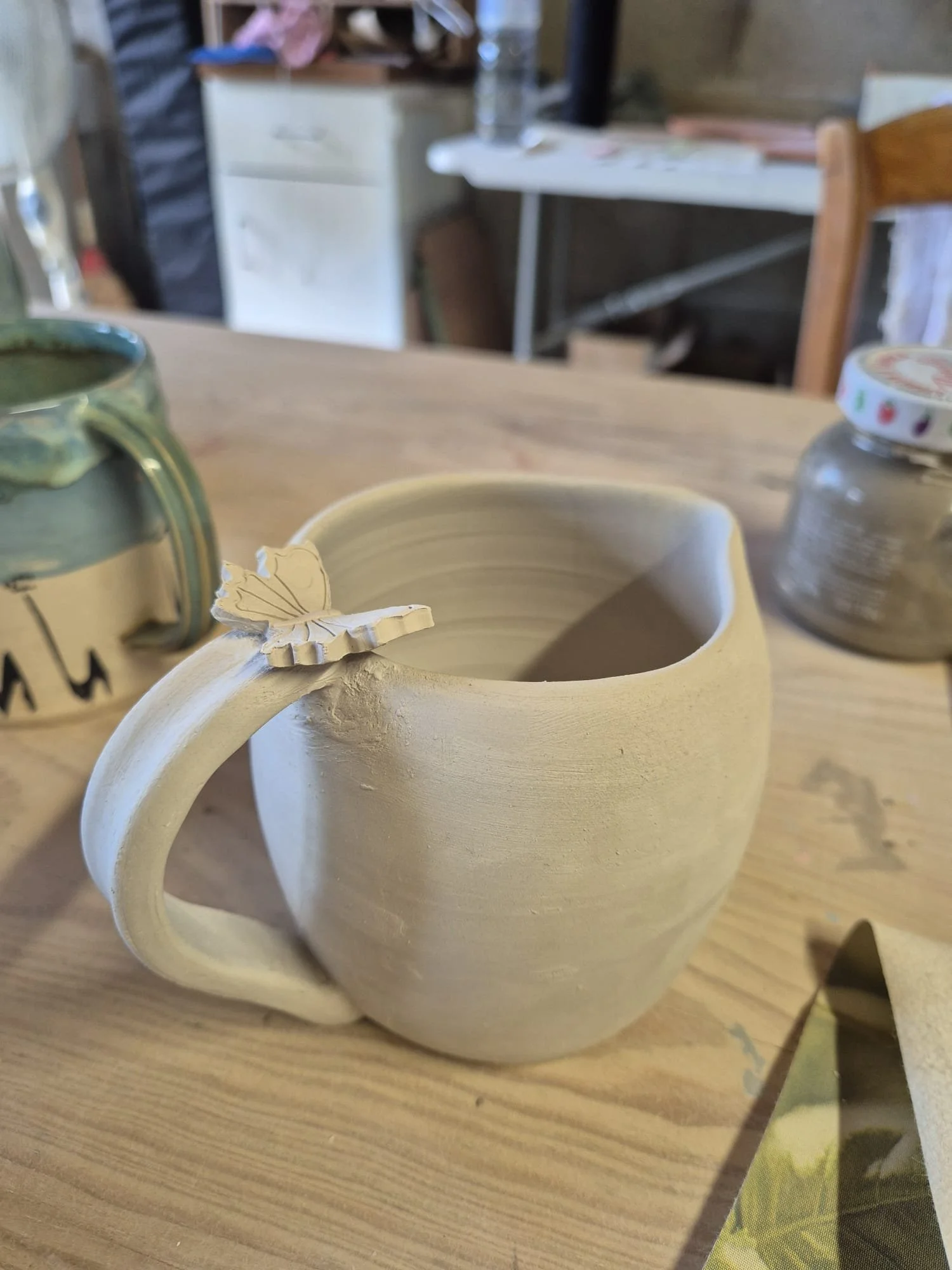 Large beige ceramic mug with a leaf-shaped decoration on a wooden table, beside a green cup and glass jar.