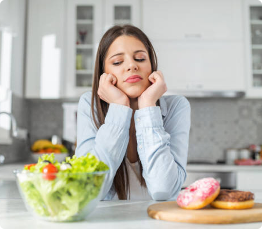 Woman resting her face in her hands beside a bowl of salad and donuts on a kitchen counter
