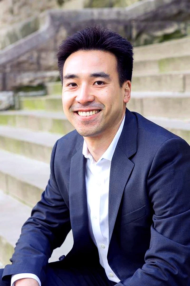 Smiling man in a navy suit and white shirt seated outdoors on concrete steps
