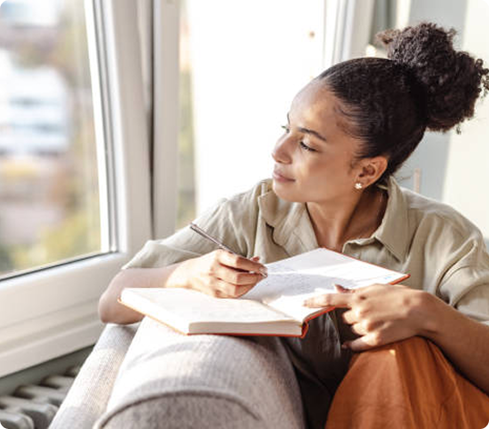 Woman with curly hair writing in a notebook while sitting by a window