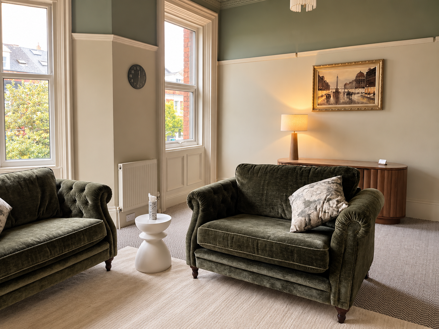 Elegant living room with green velvet sofas, white accent table, lamp, framed artwork, and large windows