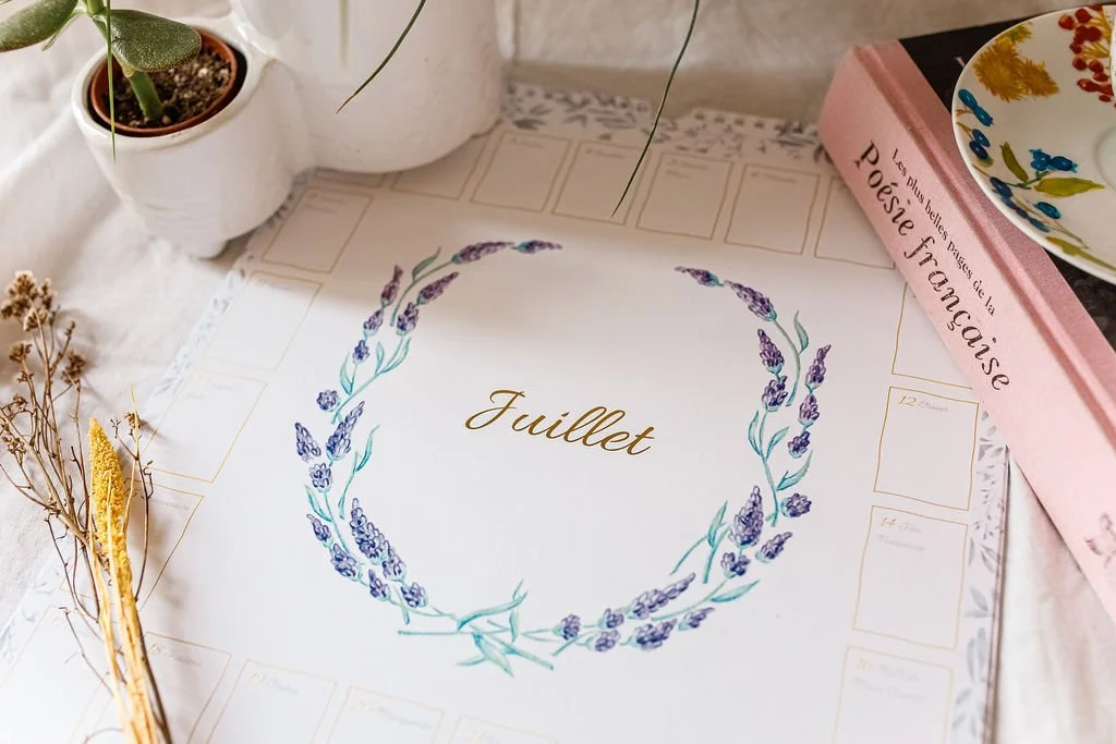 Calendrier mural de juillet décoré d’une couronne de lavande, entouré de plantes en pot, de fleurs séchées et d’un livre rose.