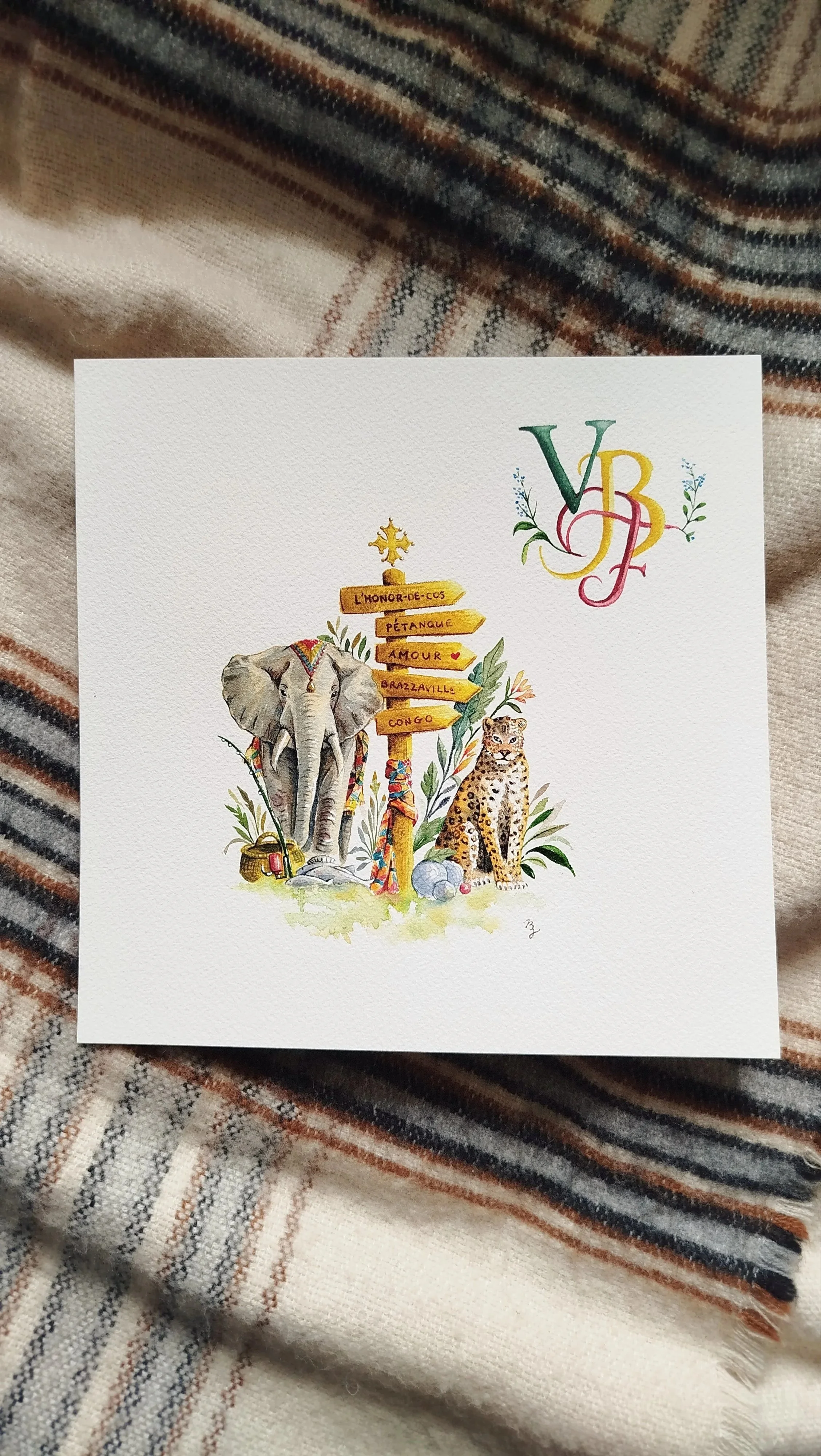 Illustration aquarelle d’un éléphant et d’un léopard entourant un panneau directionnel africain, sur une carte blanche posée sur une couverture à rayures.