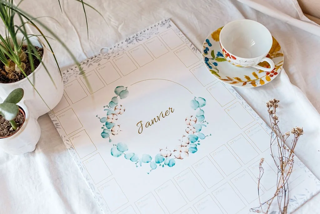 Calendrier mural illustré avec le mois de janvier, entouré de plantes en pot, d’une tasse à thé fleurie et de fleurs séchées.