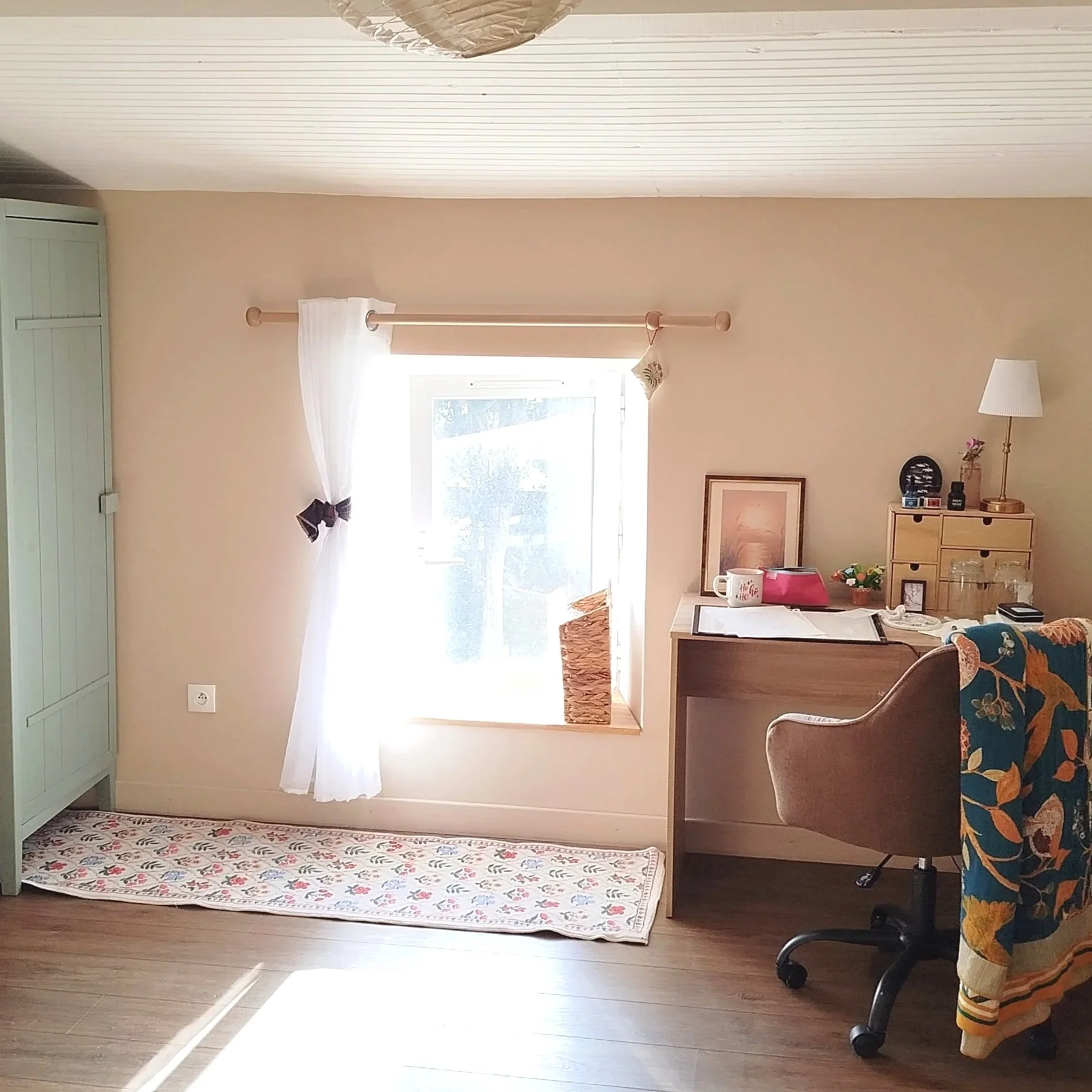 Bureau à domicile lumineux avec fenêtre, rideaux blancs, bureau en bois, lampe, chaise à roulettes et tapis fleuri.